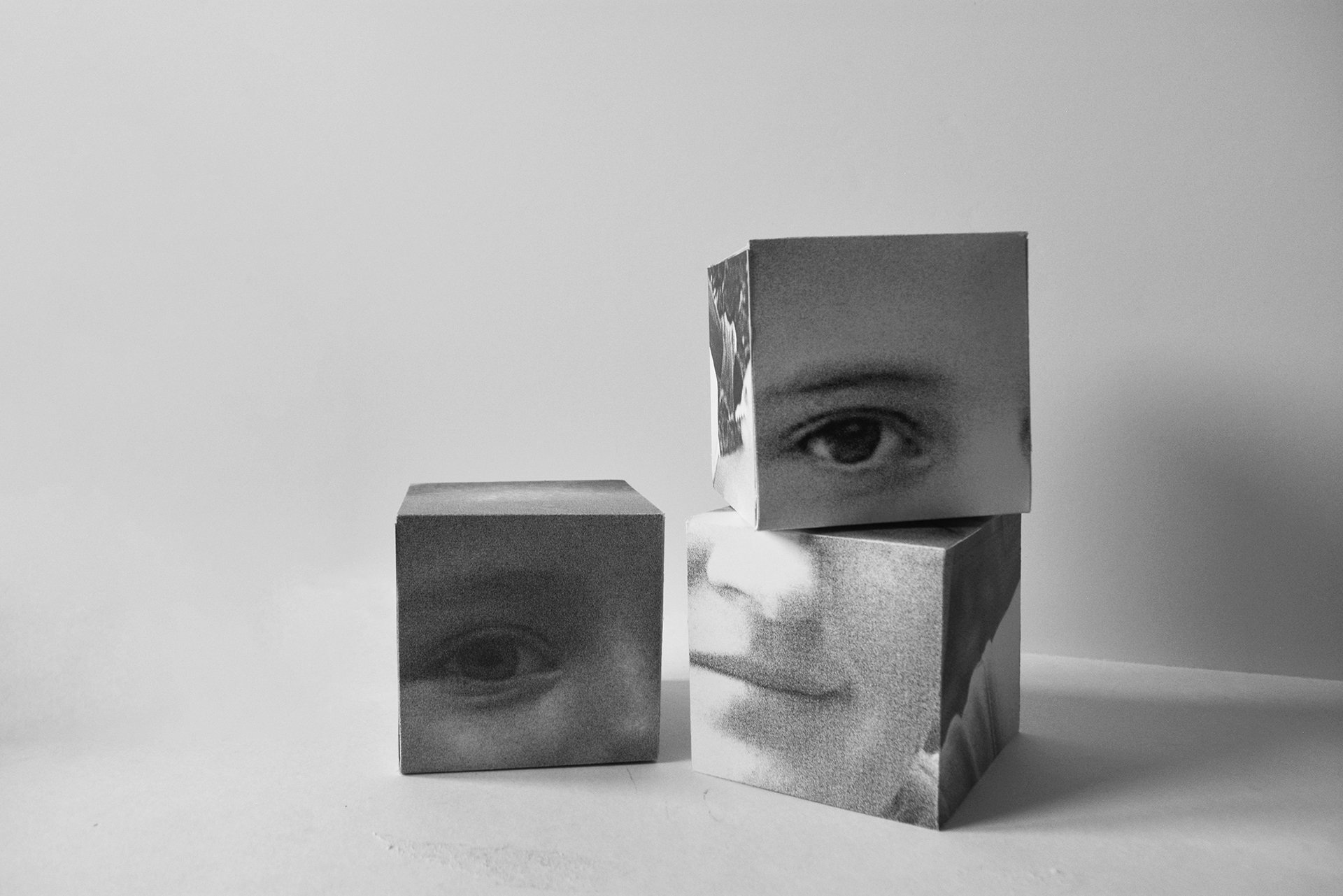 Zdjęcie 1.2 - tekst alternatywny: Fotografia trzech czarno-białych sześcianów na jasnoszarym tle, przedstawiające fragmenty różnych twarzy. Kostki ułożone są pod różnymi kątami, po prawej stronie jedna została ułożona na drugiej.