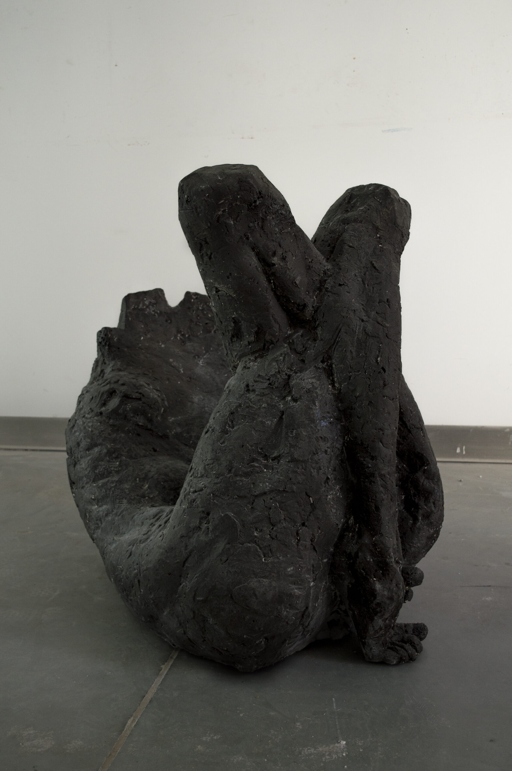 zdjęcie przedstawia ciemnoszarą rzeźbę figuratywną-skuloną postać leżącą ze skrzyżowanymi nogami. Rzeźba leży na podłodze,widok frontalny.