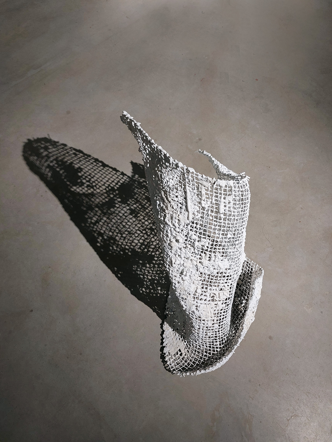Zdjęcie przedstawia fragment metalowej siatki wygiętej w taki sposób, że sugeruje ona fragment rozwianego materiału. Siatka pokryta jest białym gipsem, niektóre oczka siatki są gęsto zakryte. Na szara posadzkę siatka rzuca mocny cień, który wzmacnia ekspresję.