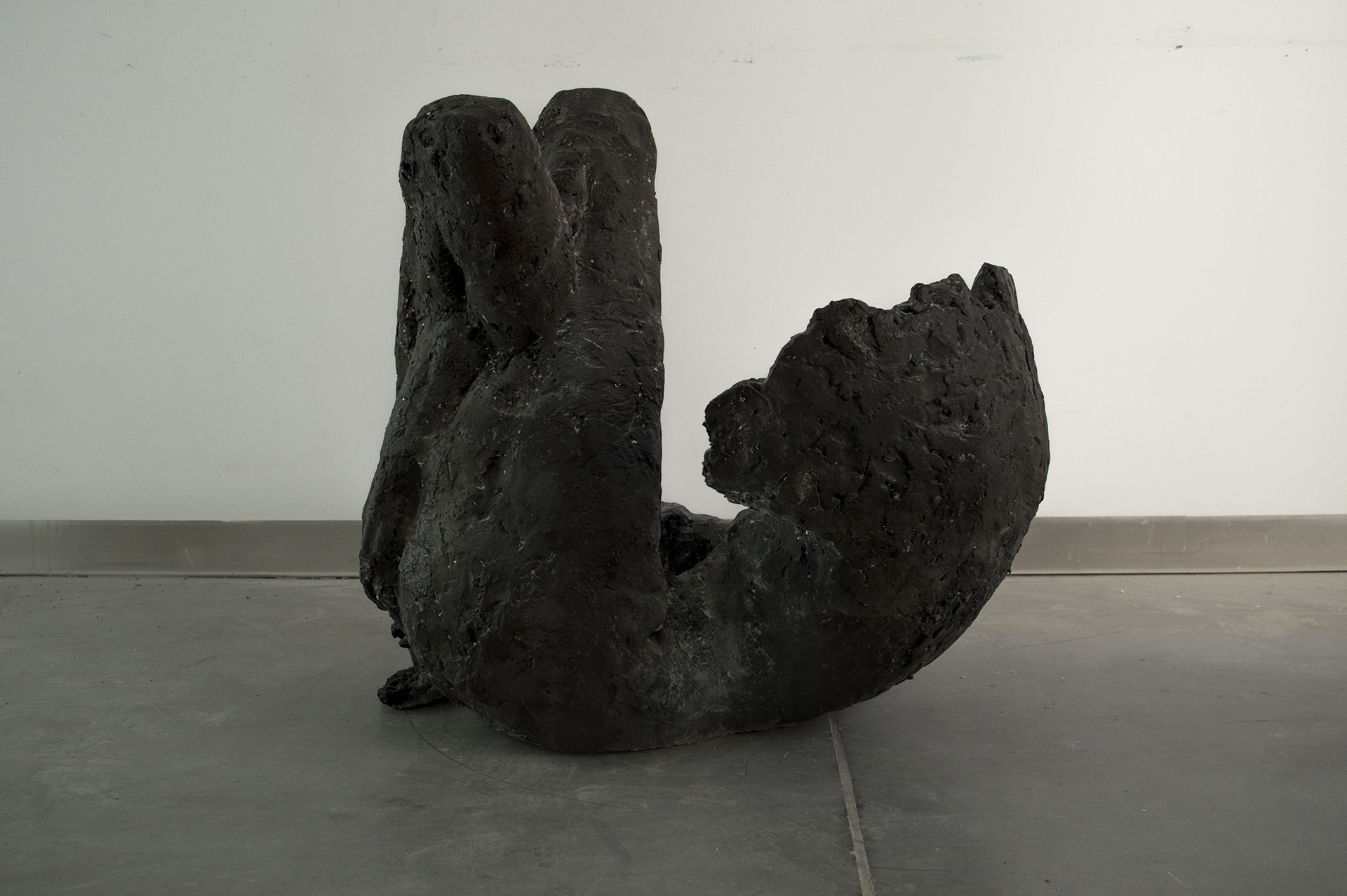 zdjęcie przedstawia ciemnoszarą rzeźbę figuratywną-skuloną postać leżącą ze skrzyżowanymi nogami. Rzeźba leży na podłodze,widok boczny.