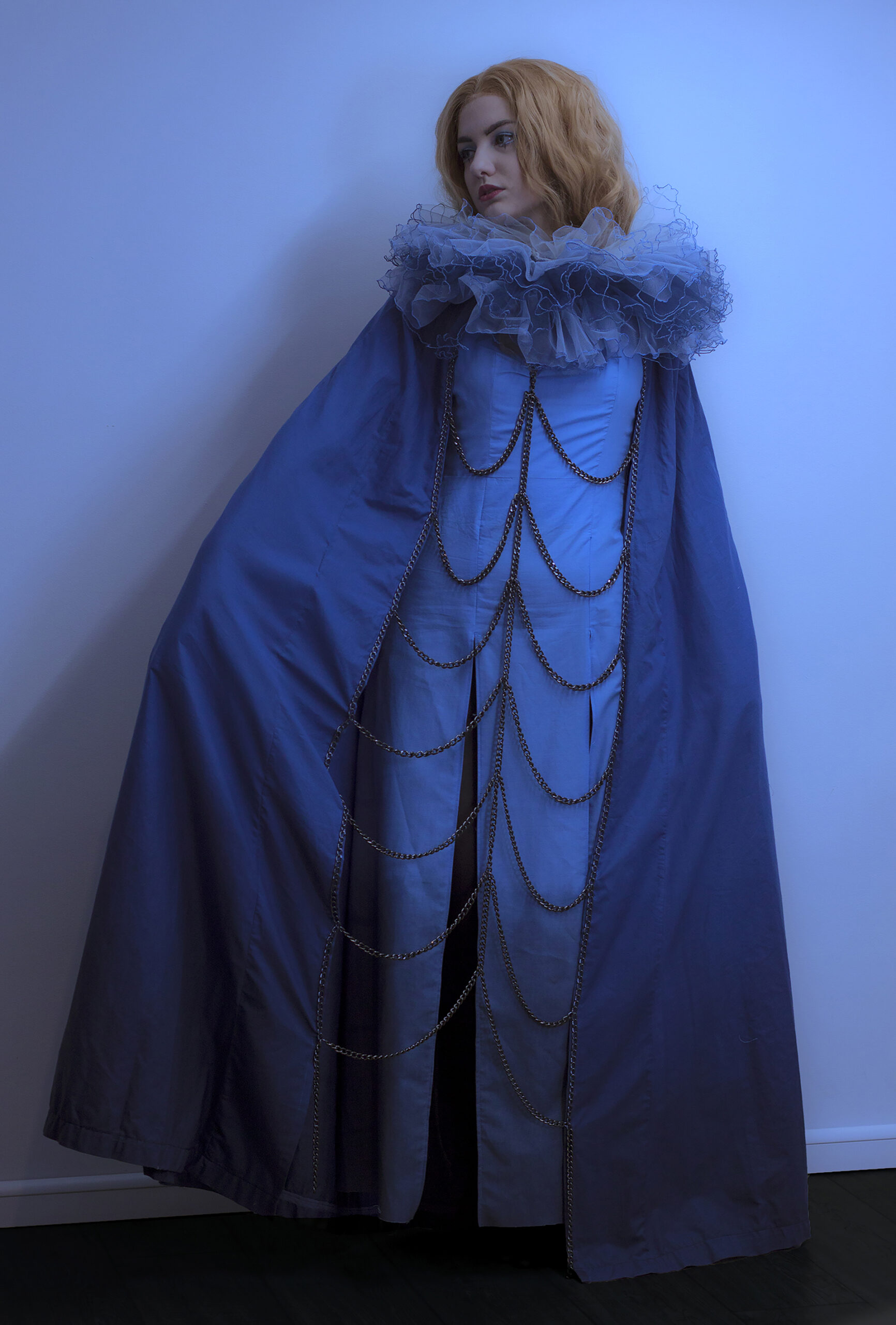 Kobieta o rudych włosach w błękitnej długiej sukience, niebieskiej pelerynie do ziemi połączonej z przodu łańcuchami i puszystą tiulową błękitną kryzą. Tło neutralne.
