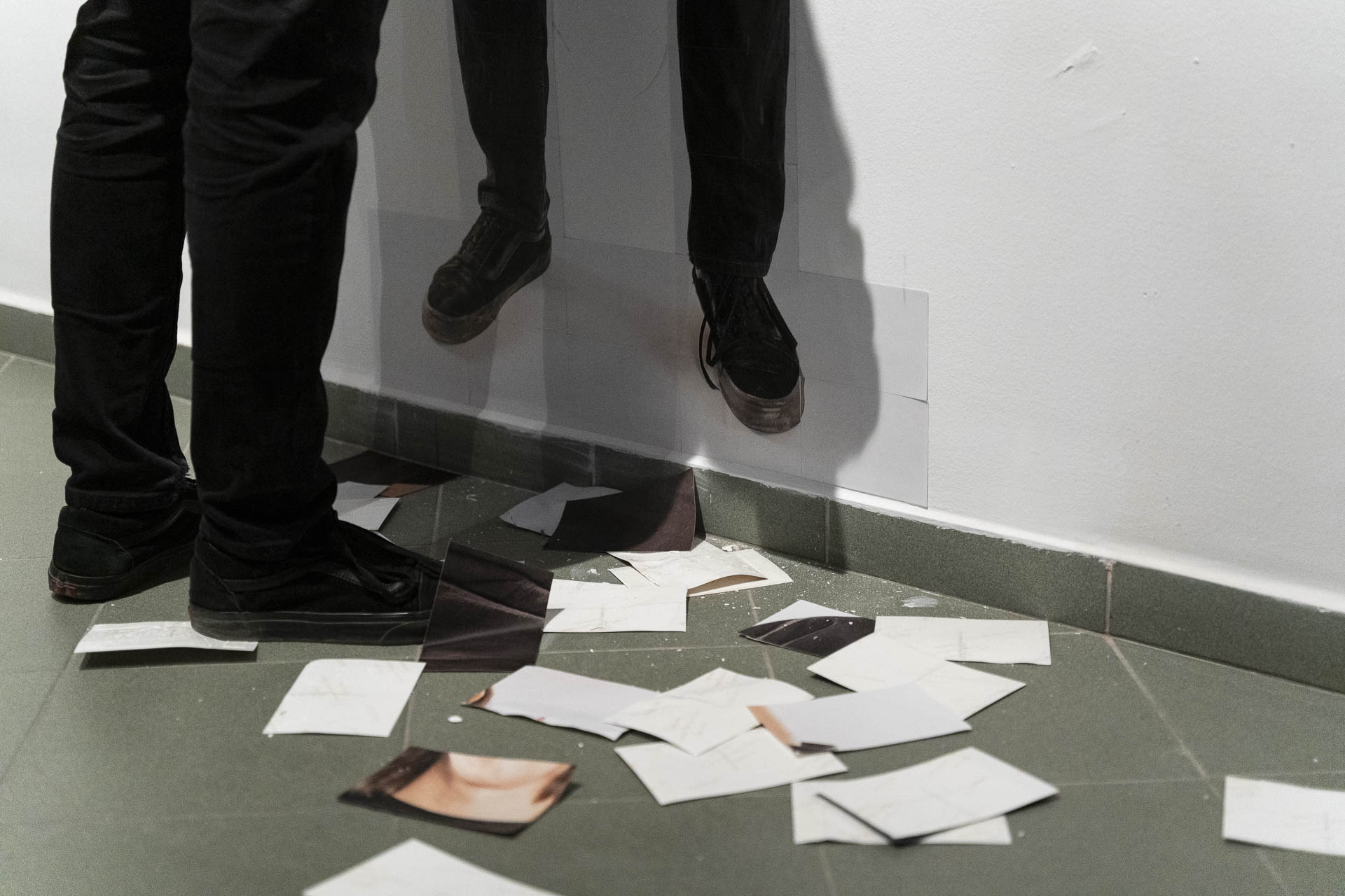 Nogi mężczyzny przy ścianie na której widać przyklejone zdjęcie tego samego fragmentu ciała w identycznych co rzeczywiste butach i spodniach. Na podłodze rozrzucone w nieładzie, wcześniej oderwane od ściany fragmenty zdjęcia