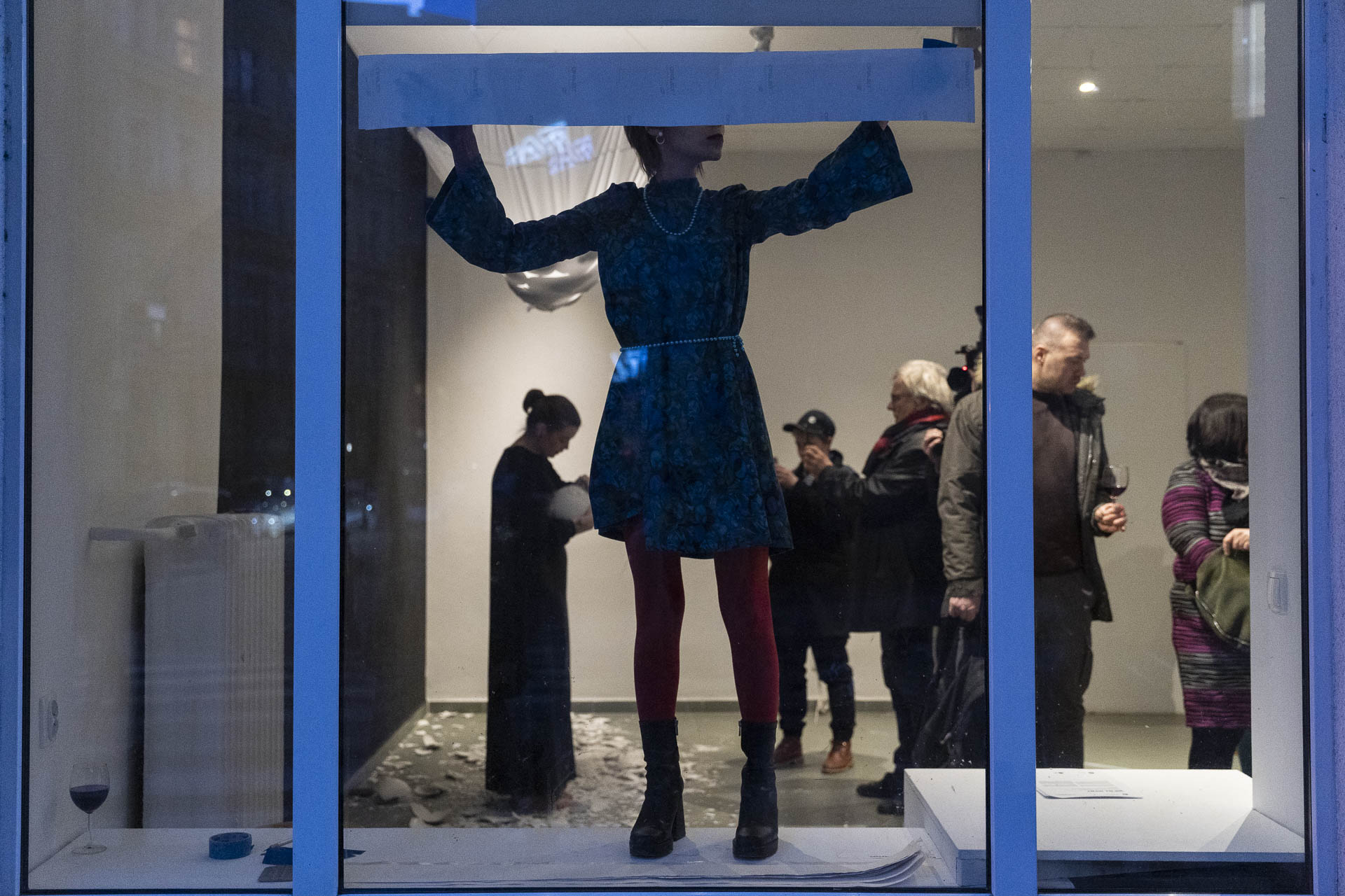 Kobieta w oknie, stojąc na parapecie, przykleja do szyby błękitno szary pas papieru na całą szerokość szyby. Za nią widoczne osoby we wnętrzu galerii.