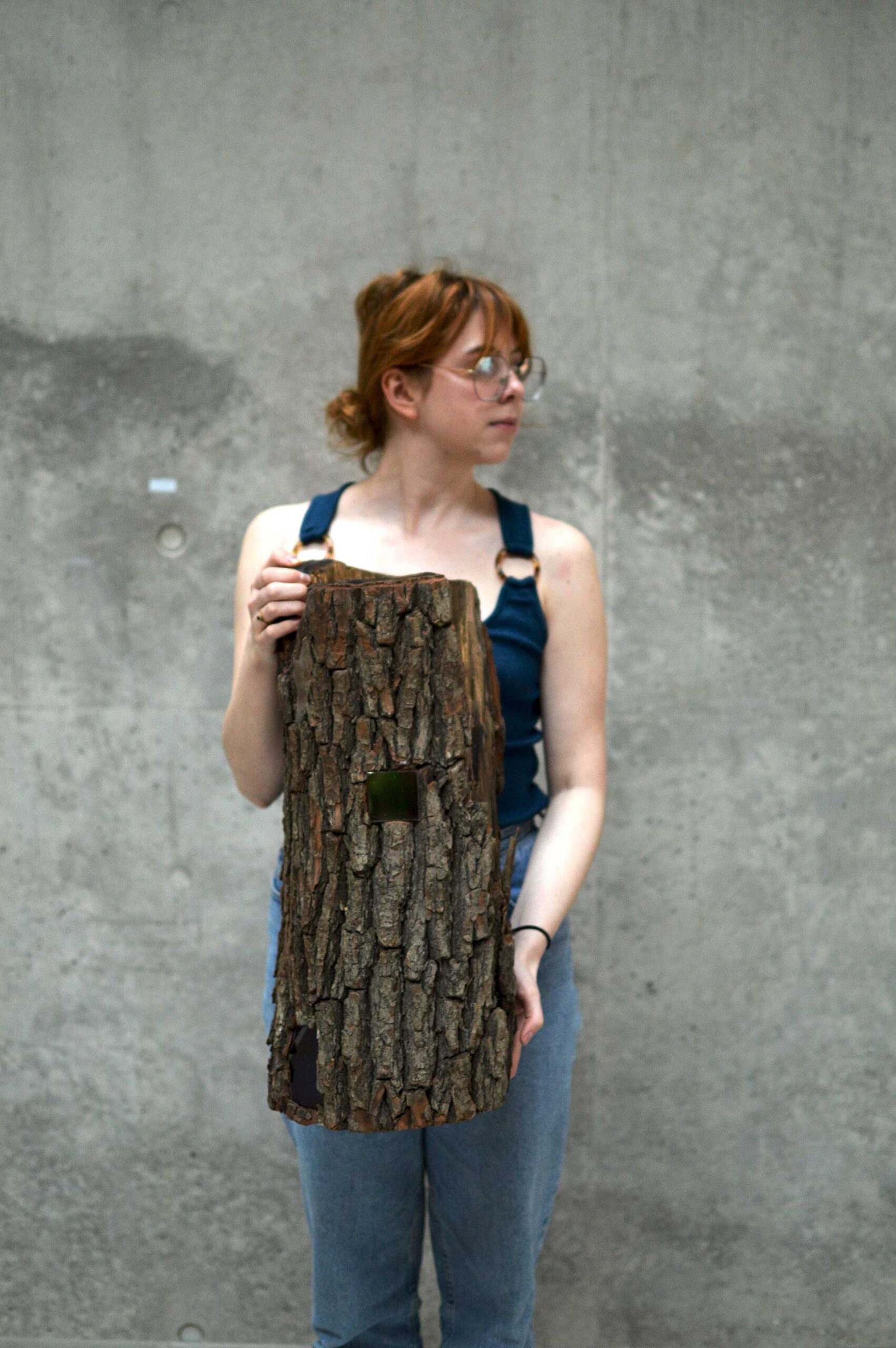 Fotografia młodej kobiety w okularach na szarym tle.Kobieta - z twarzą zwróconą w jedną stronę - w ujęciu od kolan w górę. W dłoniach trzyma pień drzewa. W tle jest betonowa ściana