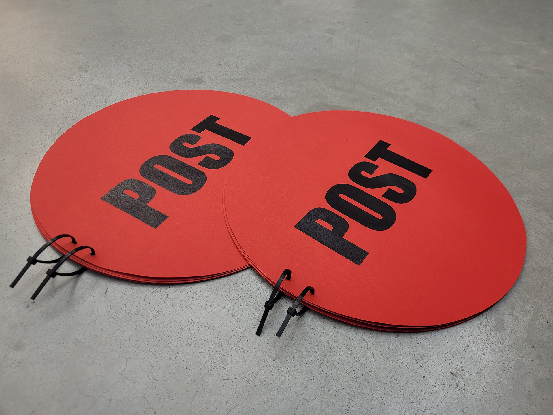 Fotografia przedstawia dwie okrągłe czerwone gazety z napisem POST, spięte trytytkami, leżą  na betonowej podłodze.