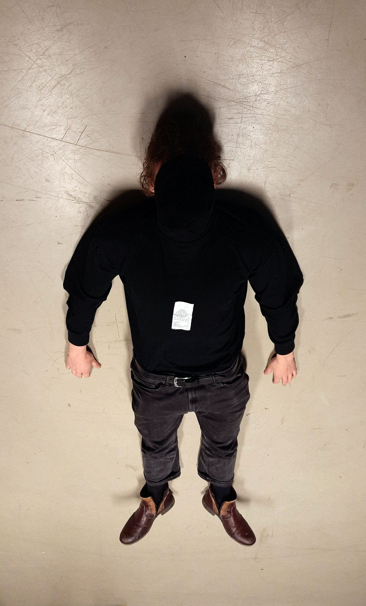 Zdjęcie przedstawia leżącą na podłodze, ubraną na czarno postać. Ma ona zakrytą twarz a na torsie położony sklepowy paragon