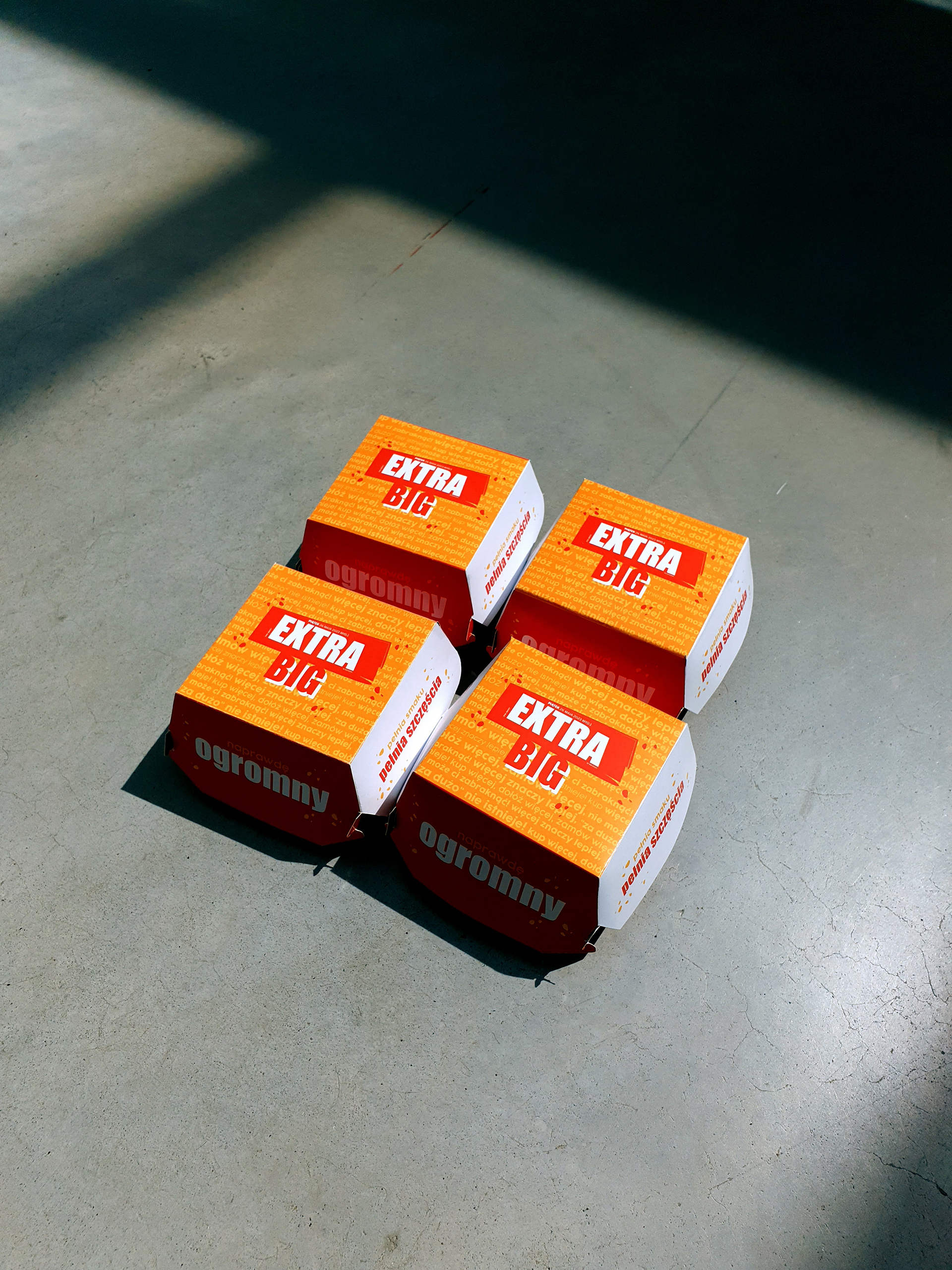 Fotografia przedstawia cztery opakowania do hamburgerów z napisem EXTRA BIG leżące na betonowej podłodze