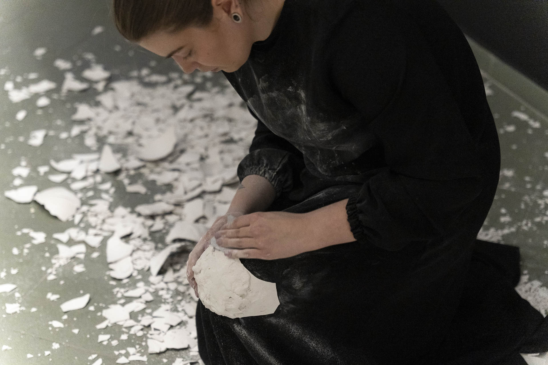 Kobieta w czarnej, zabrudzonej gipsem sukni klęczy na podłodze wśród rozbitych białych skorup. Jedną z nich trzyma w dłoniach, czyści jej krawędzie.