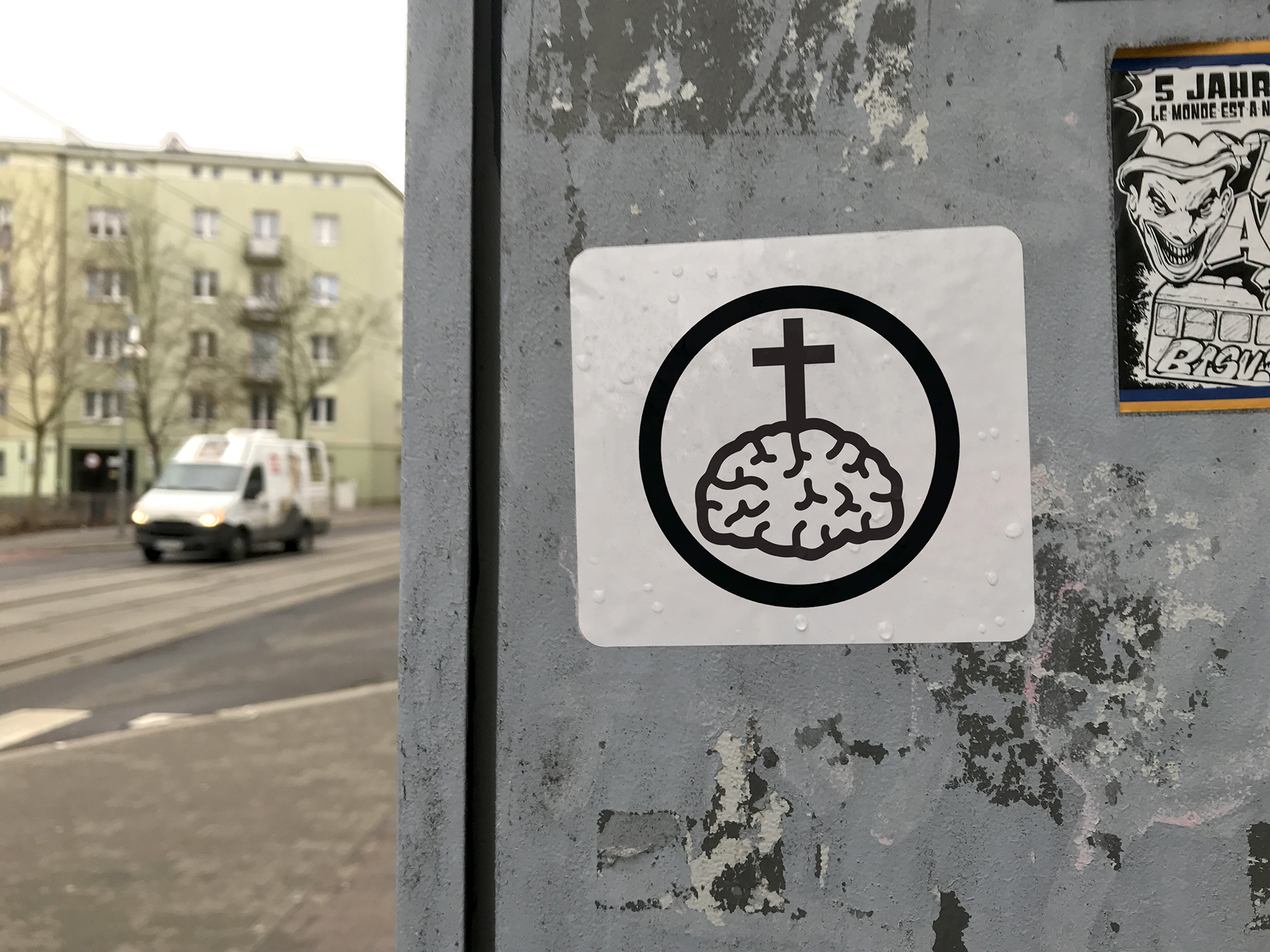 Fotografia przedstawia naklejkę w przestrzeni miejskiej na której widać zgrafizowany i przedstawiony w uproszczonej formie ludzki mózg z krzyżem, całość czarno biała wpisana w okrąg.