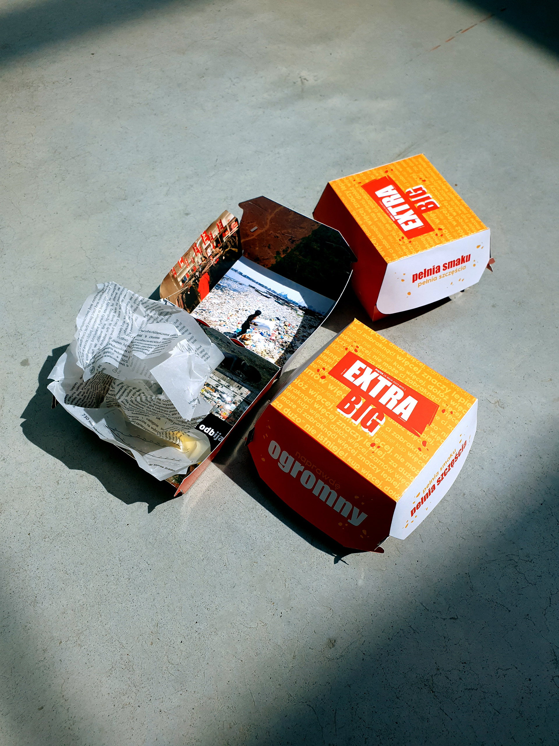 Fotografia przedstawia trzy opakowania do hamburgerów z napisem EXTRA BIG leżące na betonowej podłodze, jedno jest otwarte i znajduje się w nim zmięty papier