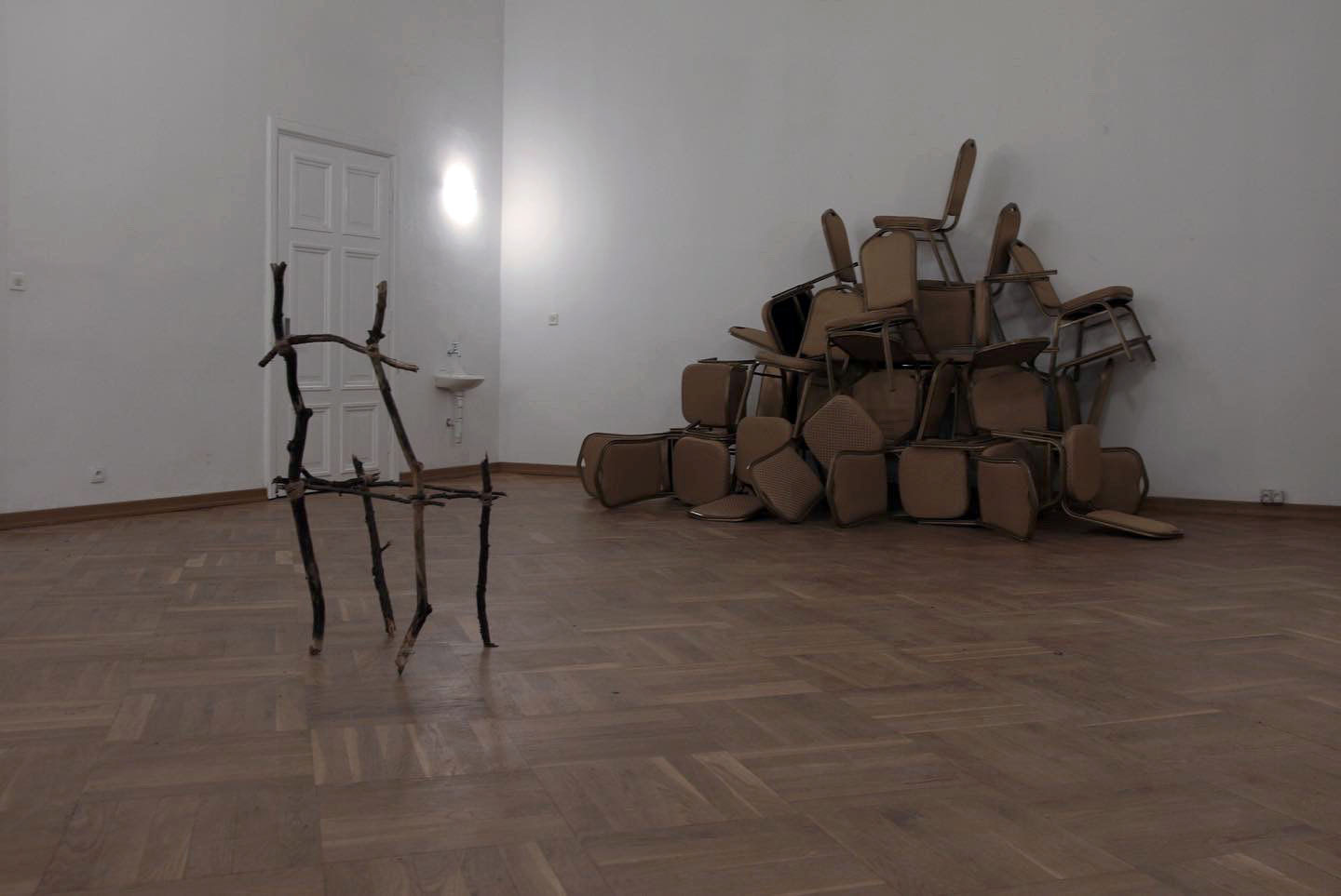 Zdjęcie przedstawia wnętrze sali. Stworzona z suchych patyków konstrukcja przypominająca puste krzesło, ustawioną jest naprzeciw stosu elegenckich krzeseł)