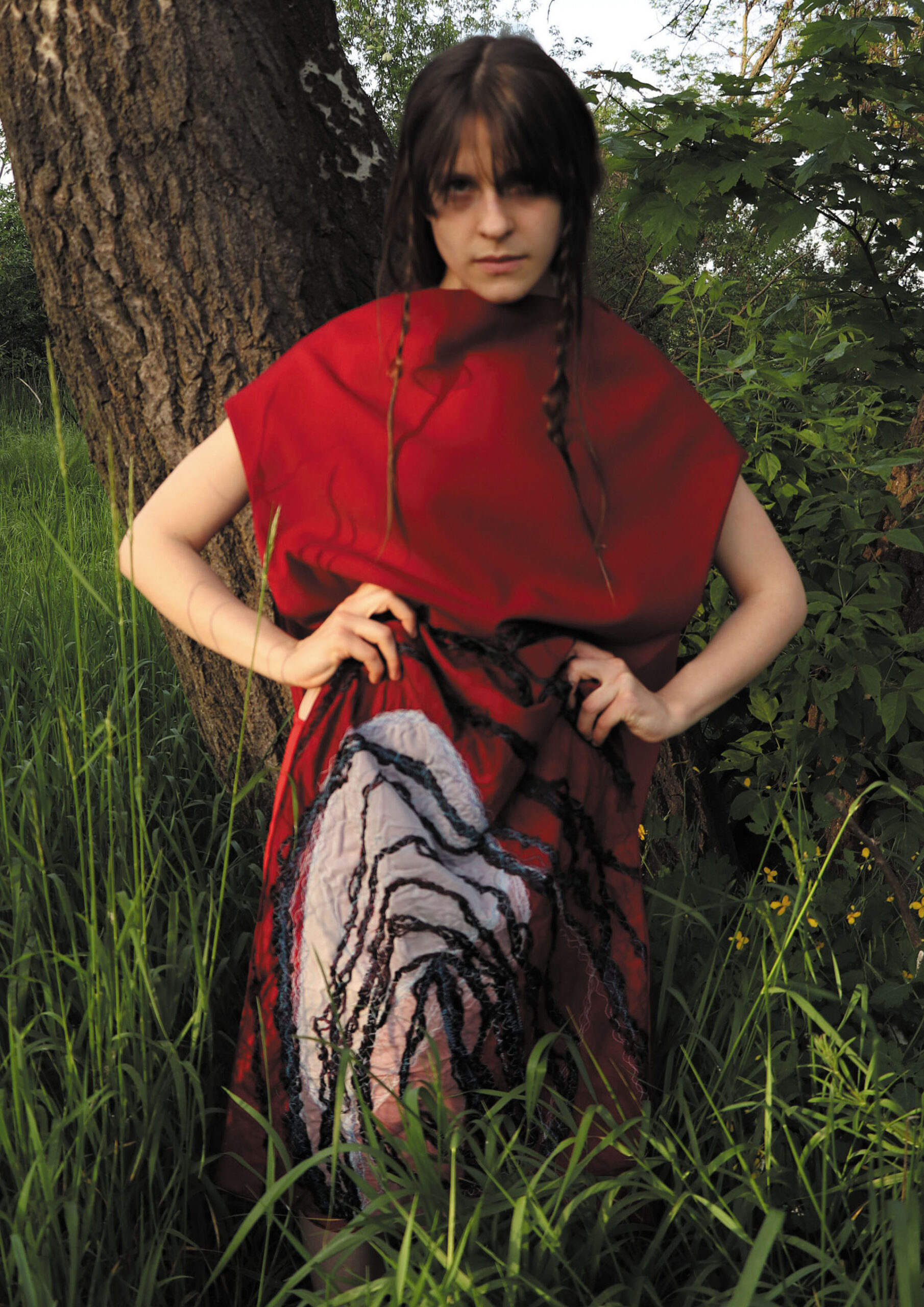 Kobieta stojąca na wprost w wysokiej trawie. Tuż za nią pień drzewa. Ubrana w czerwoną długą suknię z białym i niebieskich haftem na dolnej połowie. Długa grzywka i włosy splecione w cienkie warkocze.