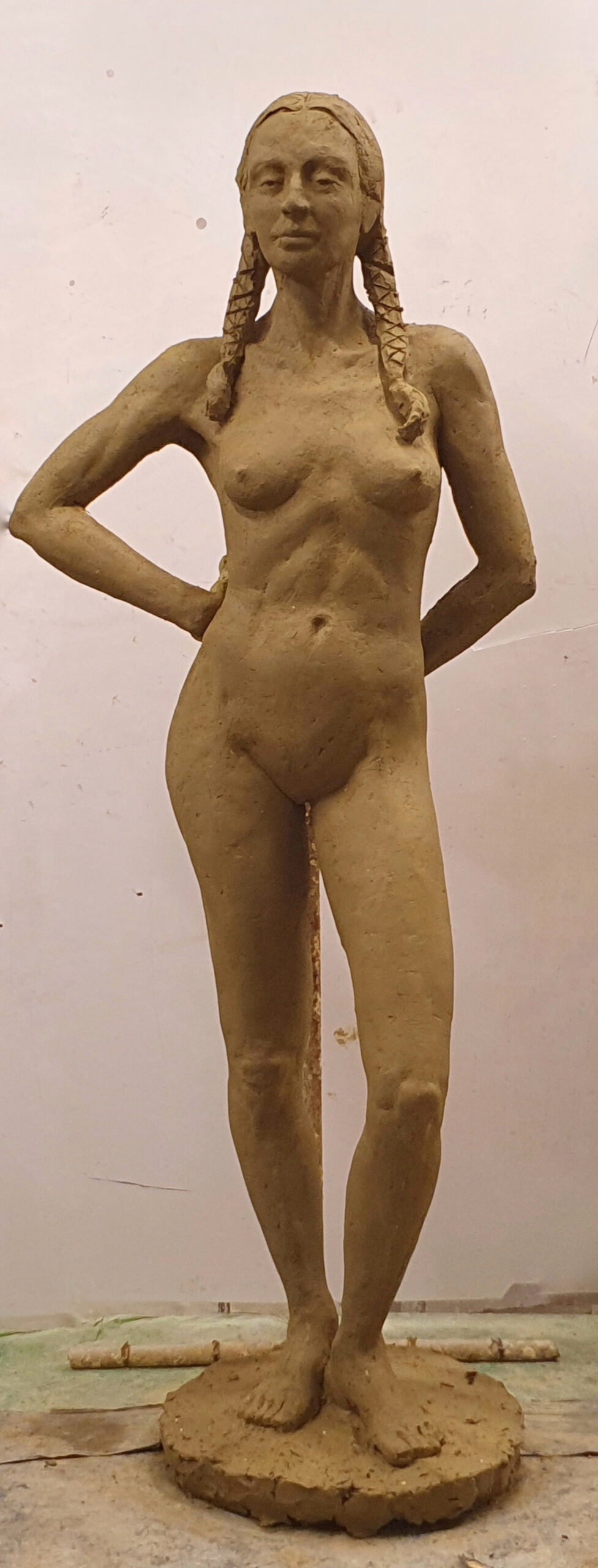 zdjęcie przedstawia rzeźbę wykonaną w glinie: postać kobiecą stojącą w kontrapoście, z ciężarem ciała na prawej nodze. Ręce ugięte w łokciach, oparte na plecach. Postać stoi na podstawie. Ujęcie frontalne