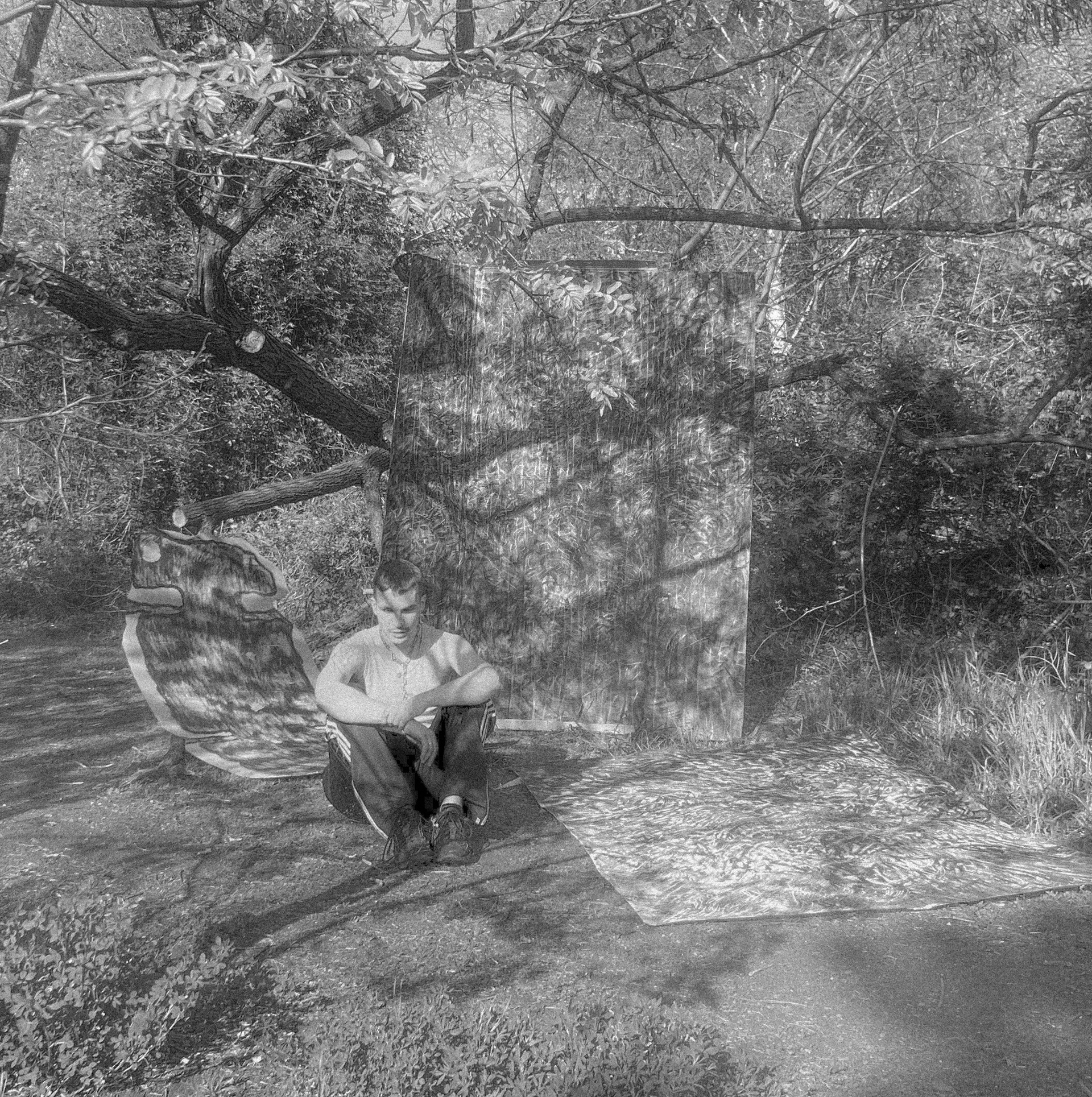 Zdjęcie Autora siedzącego pod drzewem na trawie. Obok znajdują się trzy duże rysunki na kartonach. Zdjęcie jest czarno-białe.