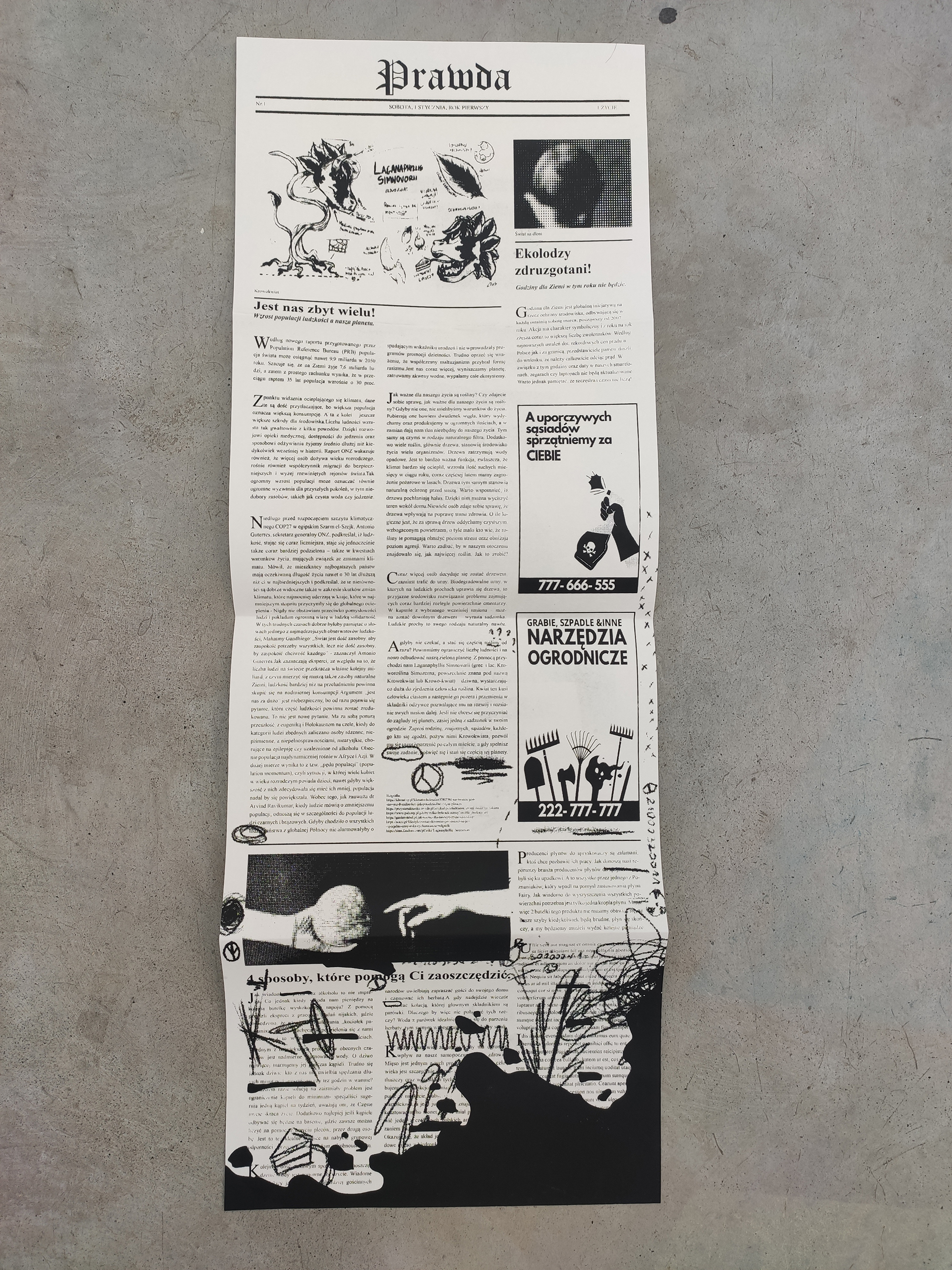 Fotografia przedstawia czarno białą gazetę w formie harmonijki rozłożoną i leżącą na betonowej podłodze.