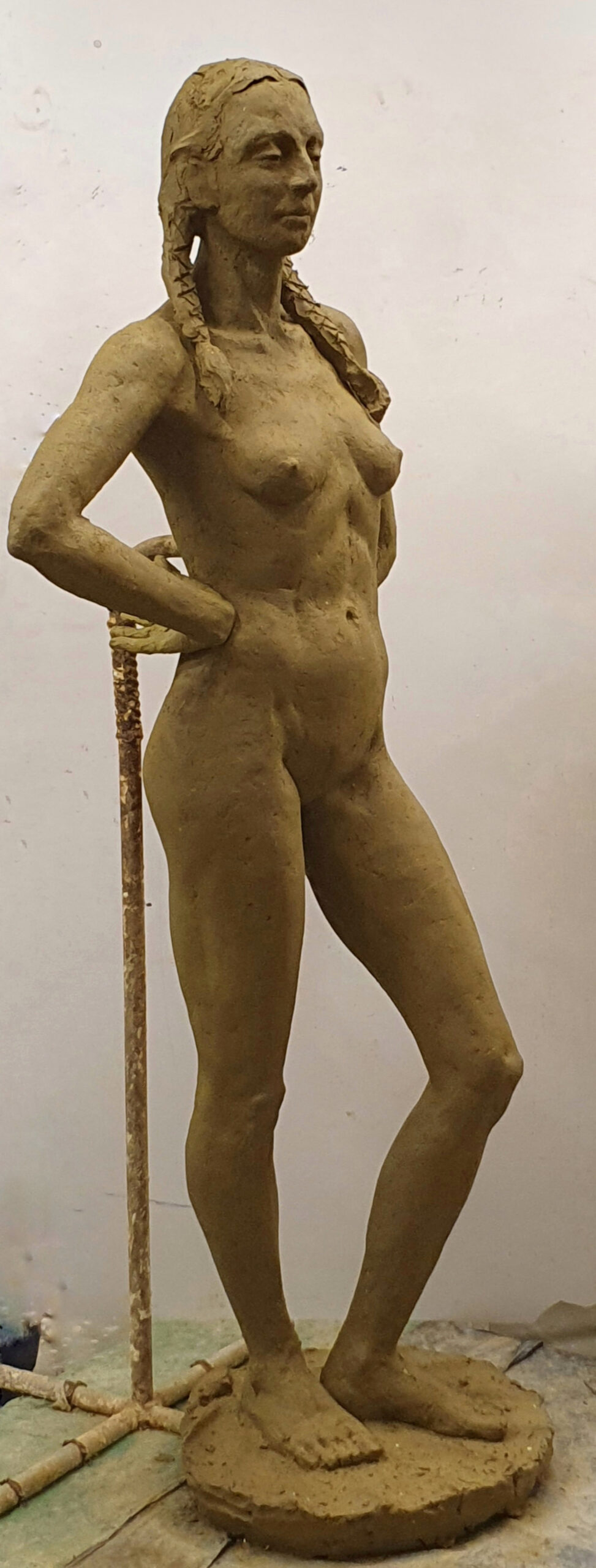  zdjęcie przedstawia rzeźbę wykonaną w glinie: postać kobiecą stojącą w kontrapoście, z ciężarem ciała na prawej nodze. Ręce ugięte w łokciach, oparte na plecach. Postać stoi na podstawie. Ujęcie boczne
