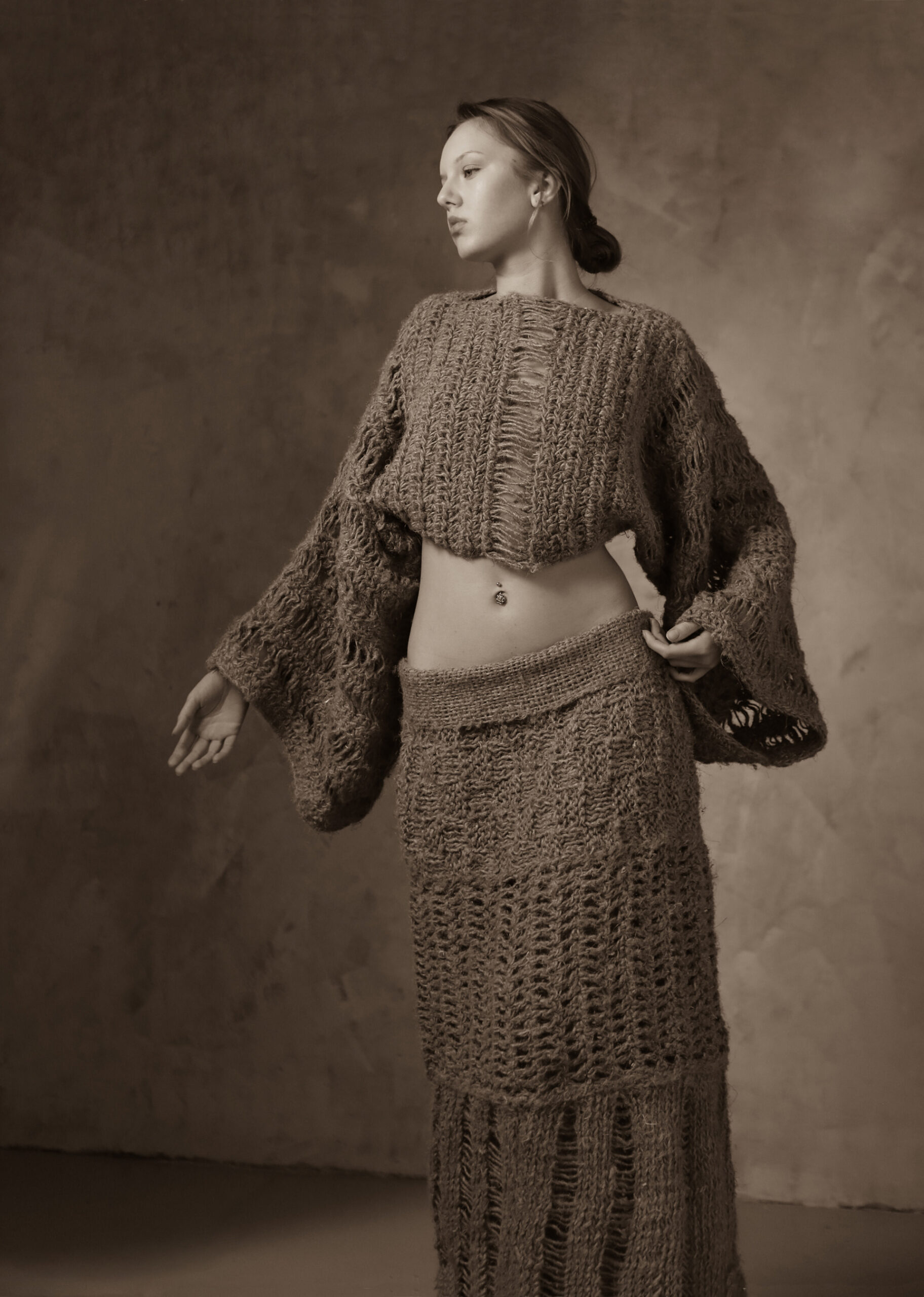 Zdjęcie w sepii. Kobieta widoczna od przodu, patrząca lekko w bok. Ubrana w komplet dziergany ze sznurka. Krótka bluzka z obfitymi długimi rękawami i długa spódnica.