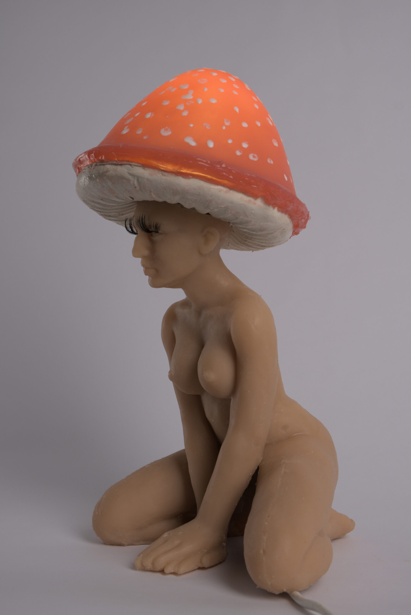 zdjęcie przedstawia lampę w formie postaci kobiecej, siedzącej na ugiętych nogach, z rękami opartymi z przodu na podłodze, skrzyżowane. Postać ma  kapelusz muchomora na głowie i sztuczne rzęsy przyklejone do powiek. Lampka jest włączona