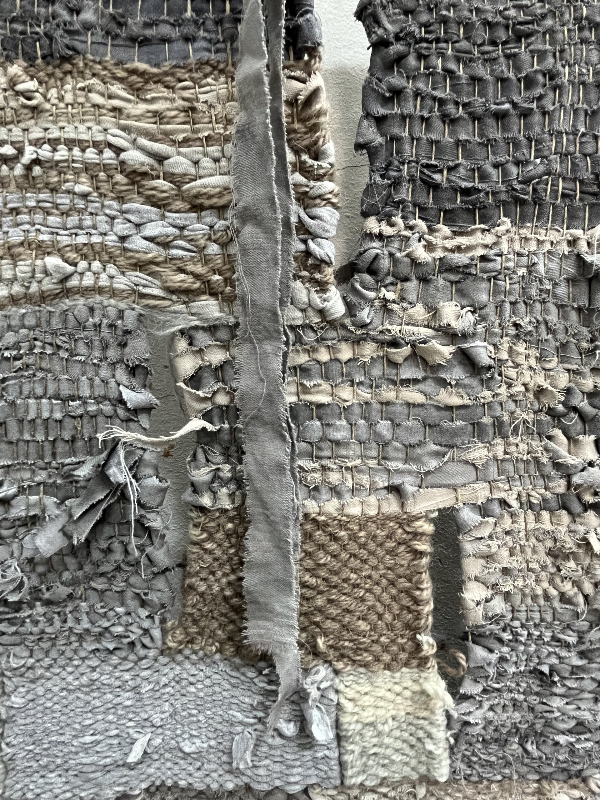 Zdjęcie przedstawia zbliżenie na tkaninę, widoczne są ażury i zwisający od góry fragment materiału wykorzystywanego przy tkaniu. Praca utrzymana w stonowanej kolorystyce.