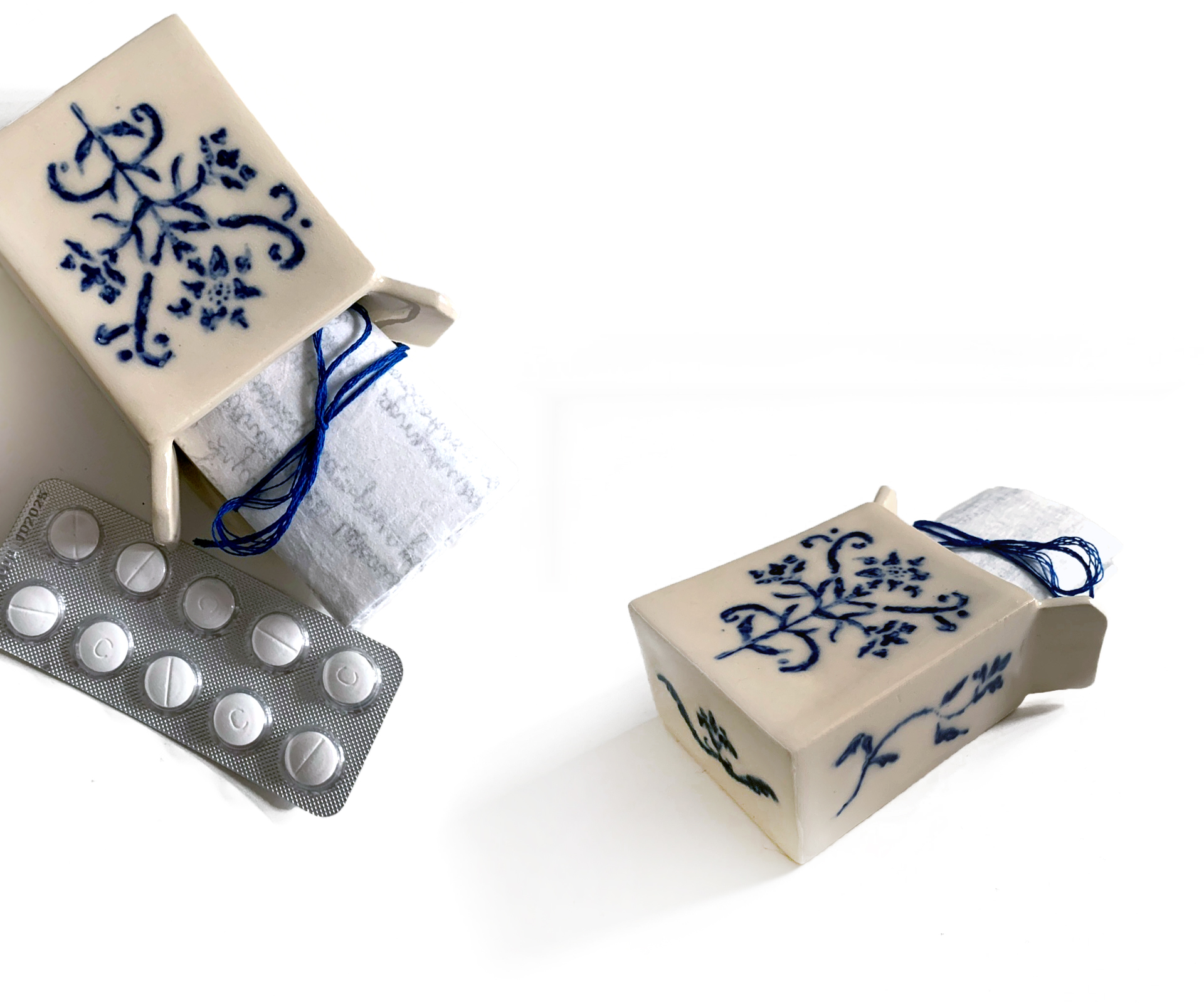 Układ z kompozycją blistra leków i pudełkami na leki wykonanymi 	z ceramiki z niebieskim wzorem. Z pudełek wystaje zwinięty 	zapisany papier przewiązany niebieską nicią.