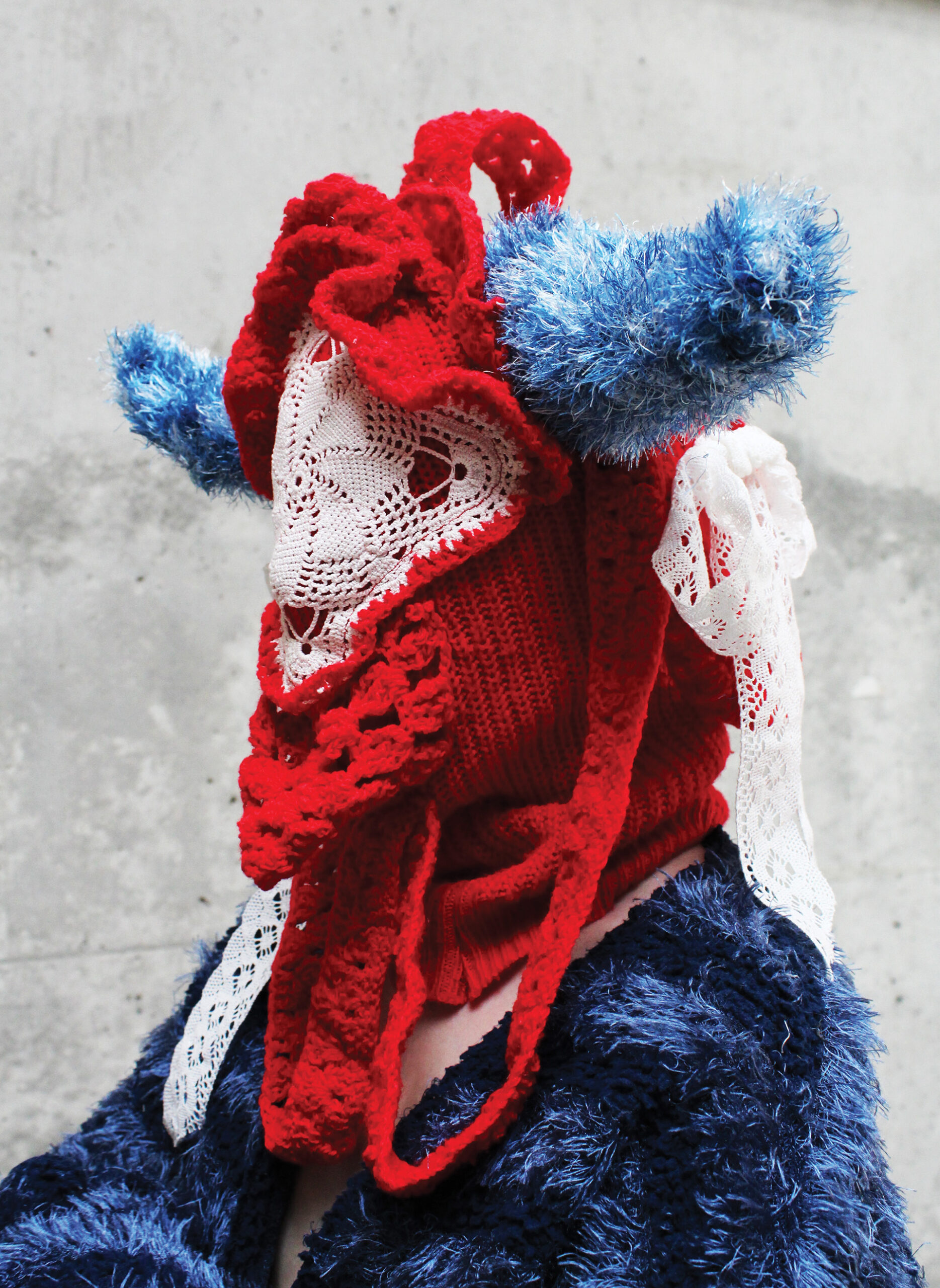 Portret człowieka w formie zakrywającej twarz, głowę i ramiona. Czerwona kominiarka z białą serwetą na twarzy i niebieskimi rogami. Na ramionach niebieski sweter w granatowe pasy.