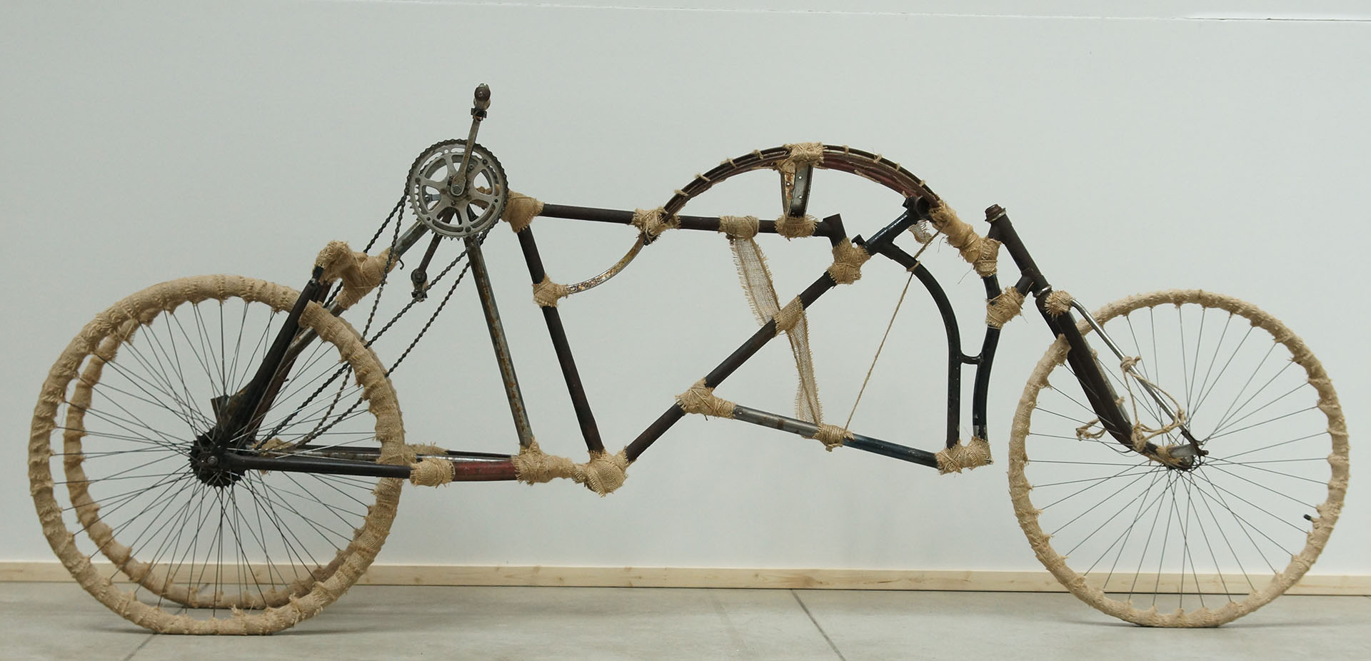 Zdjęcie przedstawia konstrukcję roweru stworzoną z pięciu ram rowerowych. Obiekt nawiązuje do nietypowych i kontrowersyjnych konstrukcji z filmu Mad Max. Użytkownik korzystający z roweru jest w założeniu projektu w pozycji leżącej. Obiekt napędzany jest siłą ludzkich rąk, korby zębate umieszczone po obu stronach suportu przenoszą napęd na tylne koła znajdujące się z przodu. Skrętne jest przednie koło znajdujące się w tylnej części pojazdu, użytkownik wykonuje skręty nogami. Łączenia ram zakrywają detale w postaci sznurków i taśmy jutowej. Koła nie posiadają już opon, zostały zastąpione taśmą jutową dla wzbogacenia detalu
