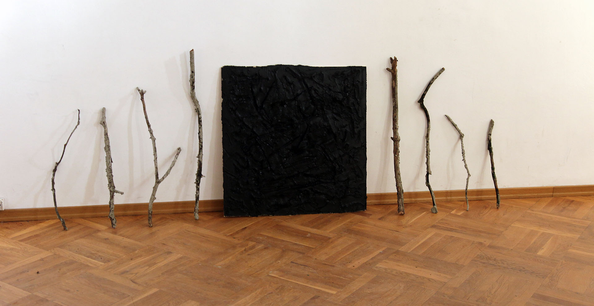 Zdjęcie przedstawia oparte o ścianę stare gałęzie, długości około 1 metra oraz kwadratową płytę o podobnych wymiarach z nałożoną czarną, smolistą substancją