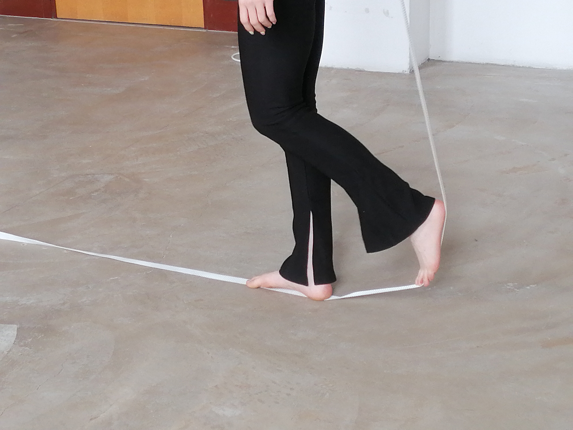 Kadr na którym widoczne są nagie stopy kształtujące przebieg białej taśmy w ten sposób że jedna przyciska ją do podłogi, druga ustawiona prostopadle napina ją do tyłu