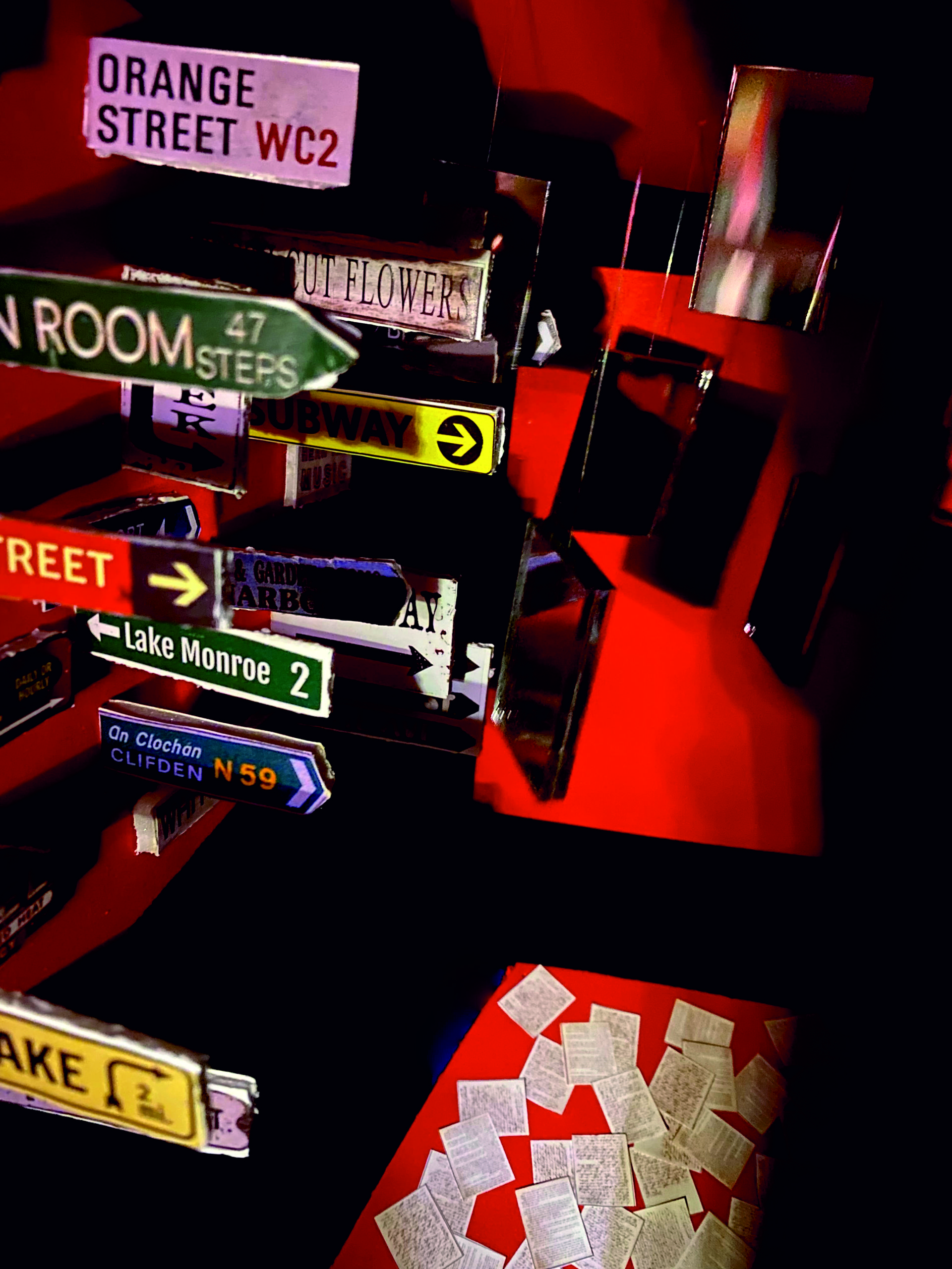 Czerwono-czarna przestrzeń wypełniona lustrami, kartkami z gazet i znakami drogowymi. W środku postać.