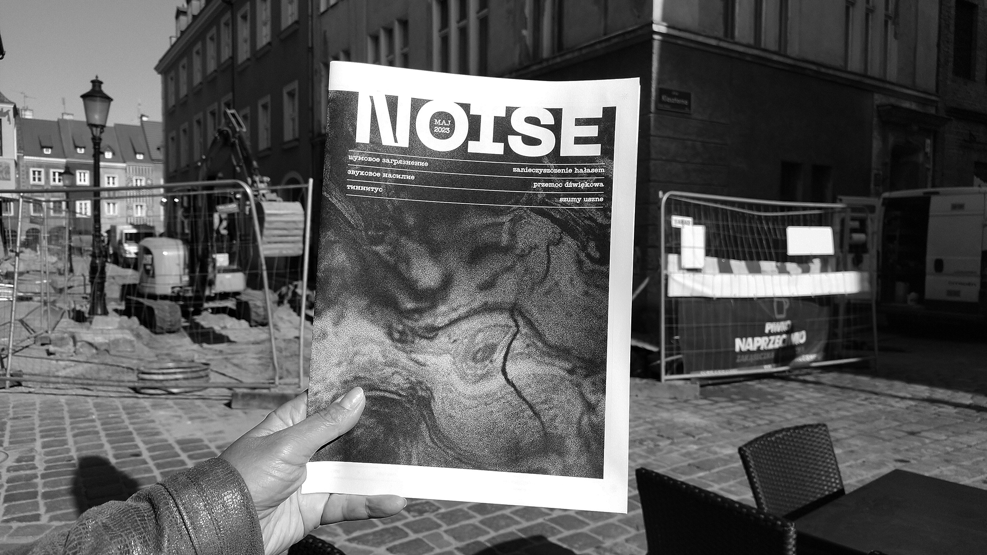Fotografia przedstawia gazetę o tytule Noise trzymaną w ręce. W tle ulice miasta podczas remontu nawierzchni, widoczne maszyny - koparka i wykopy. Fotografia czarno-biała.