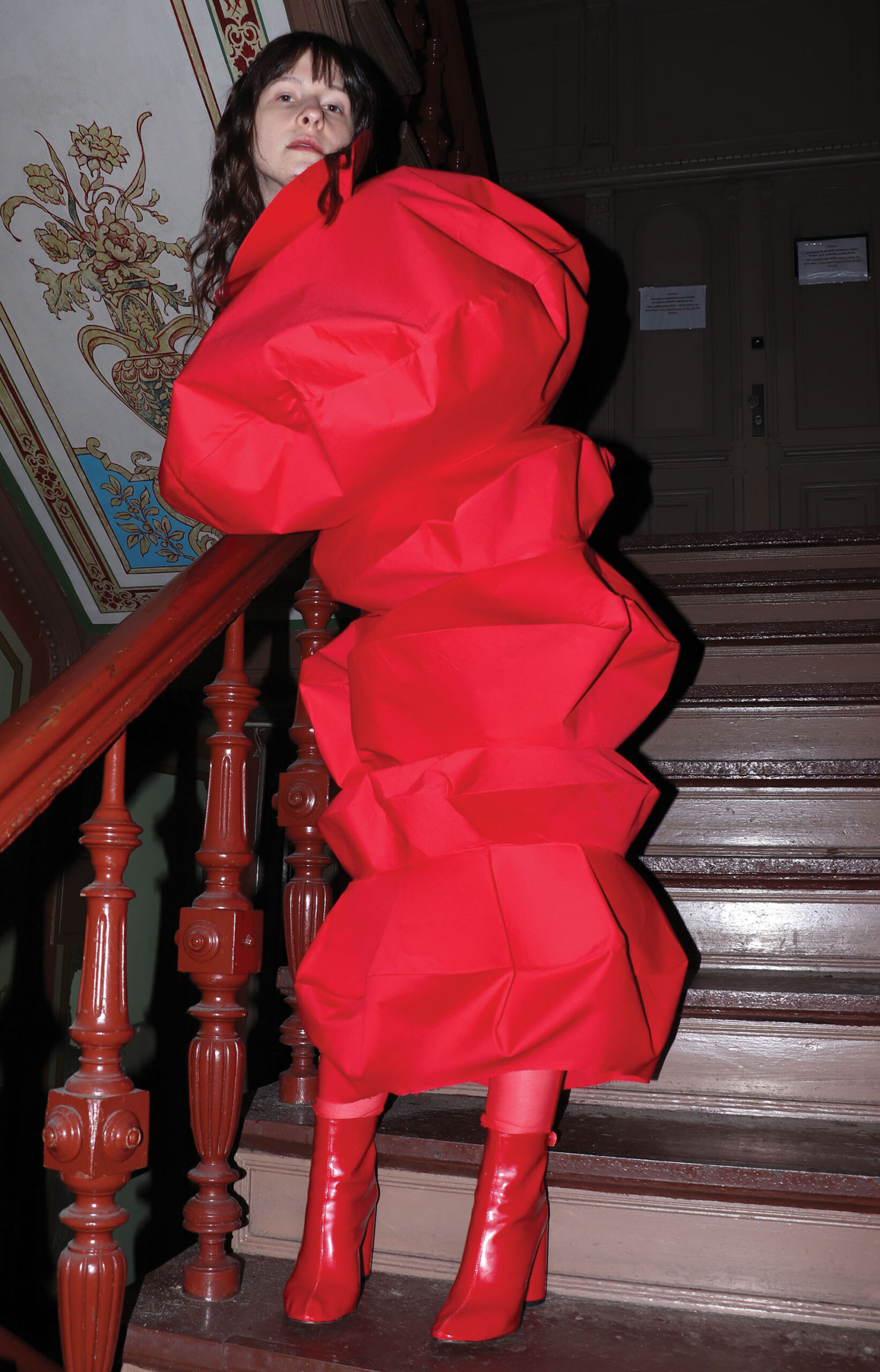 Kobieta stojąca na wprost na klatce schodowej w starej kamienicy. Ubrana w czerwoną abstrakcyjną formę zakrywającą ciało od szyi po łydki i czerwone buty na obcasie.