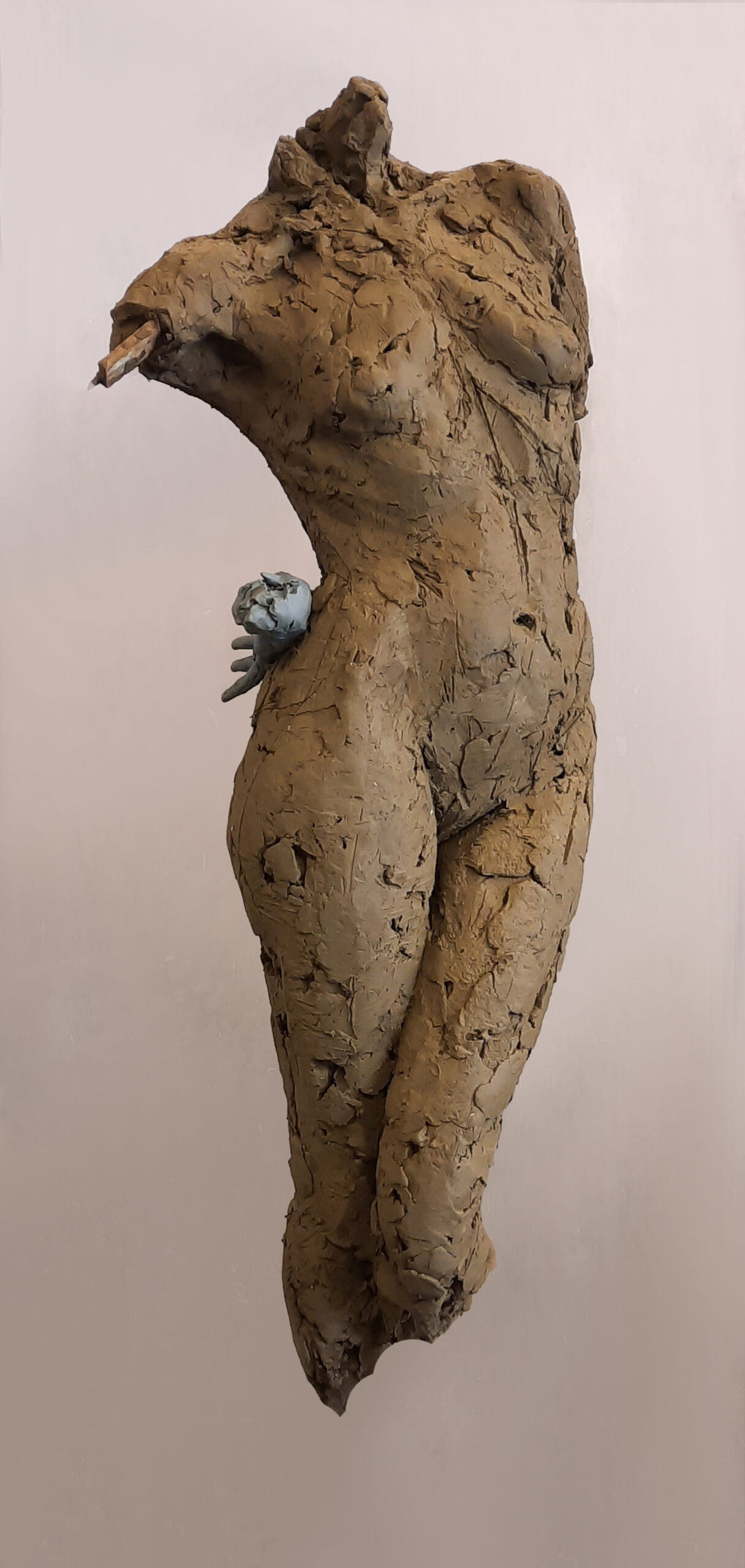 zdjęcie przedstawia rzeźbę z gliny- postać kobiecą w ujęciu od szyi do kolan, bez rąk,  jedna noga lekko ugięta. Struktura rzeźby fakturalna,ujęcie boczne