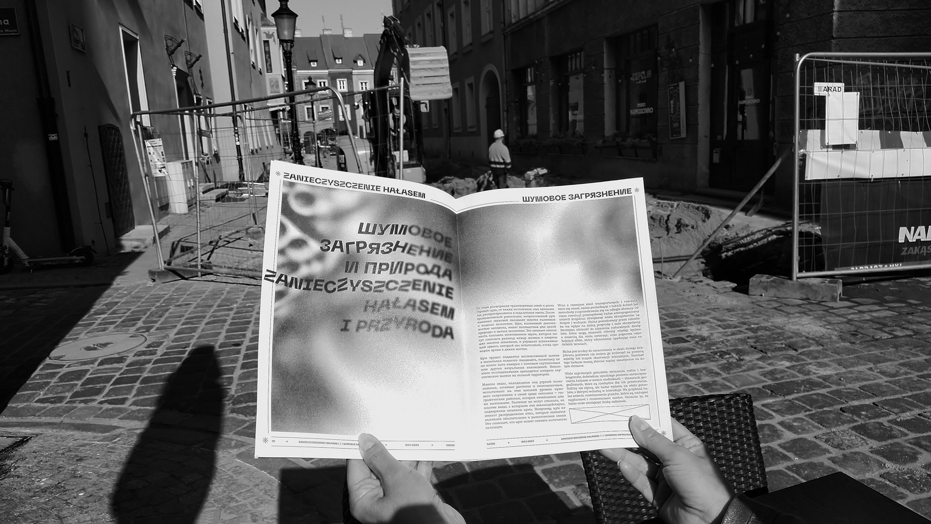 Fotografia przedstawia rozłożoną czarno białą gazetę trzymaną w rękach. W tle ulice miasta podczas remontu nawierzchni, widoczne maszyny - koparka i wykopy. Fotografia czarno-biała.