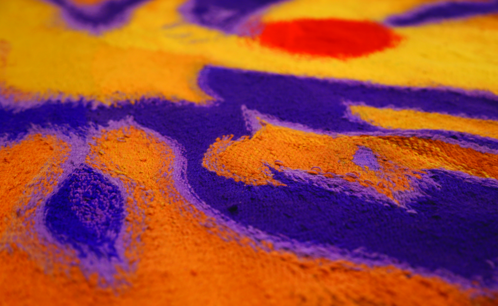  Zdjęcie przedstawia fragment tkaniny, wykonanej za pomocą drobnego wątku  na której rozgrywa się wielobarwna abstrakcja w ciepłych tonacjach