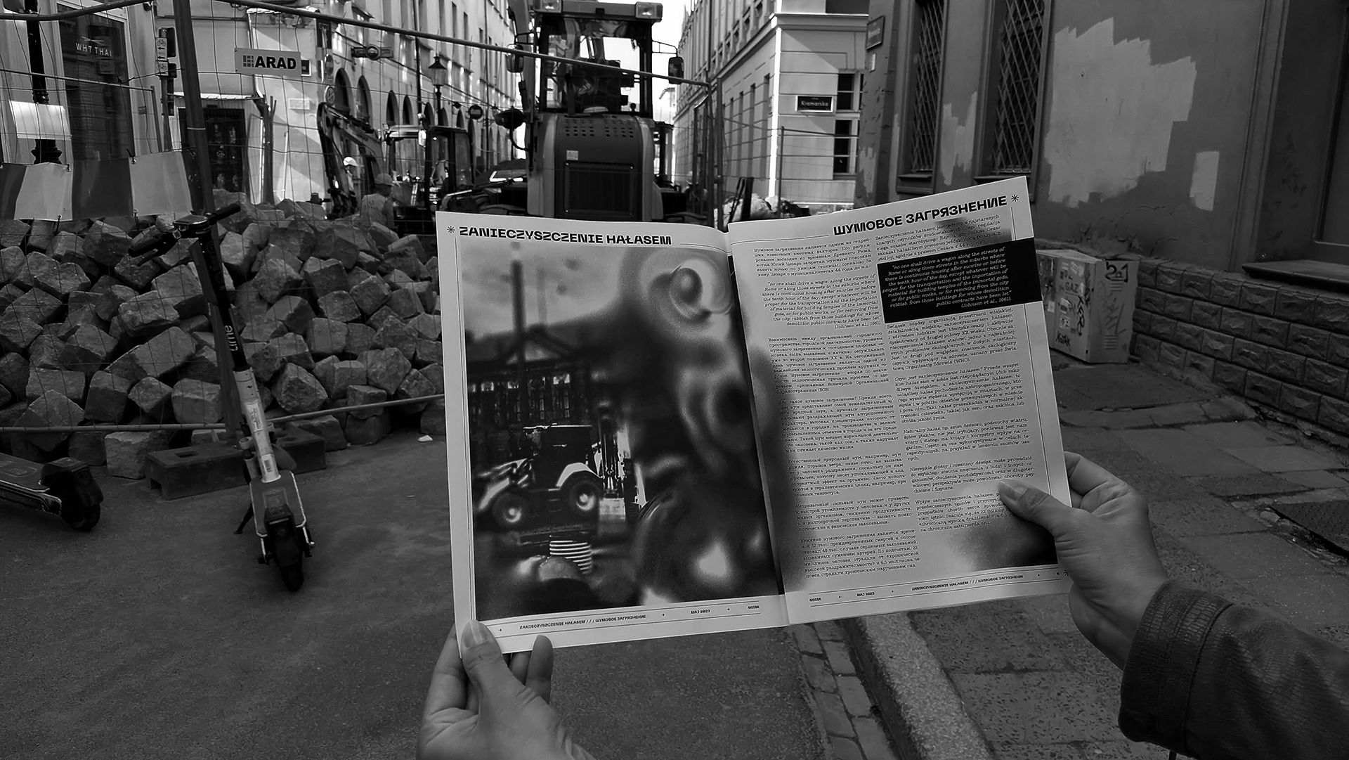 Fotografia przedstawia rozłożoną czarno białą gazetę trzymaną w rękach. W tle ulice miasta podczas remontu nawierzchni, widoczne maszyny - koparka i wykopy. Fotografia czarno-biała.