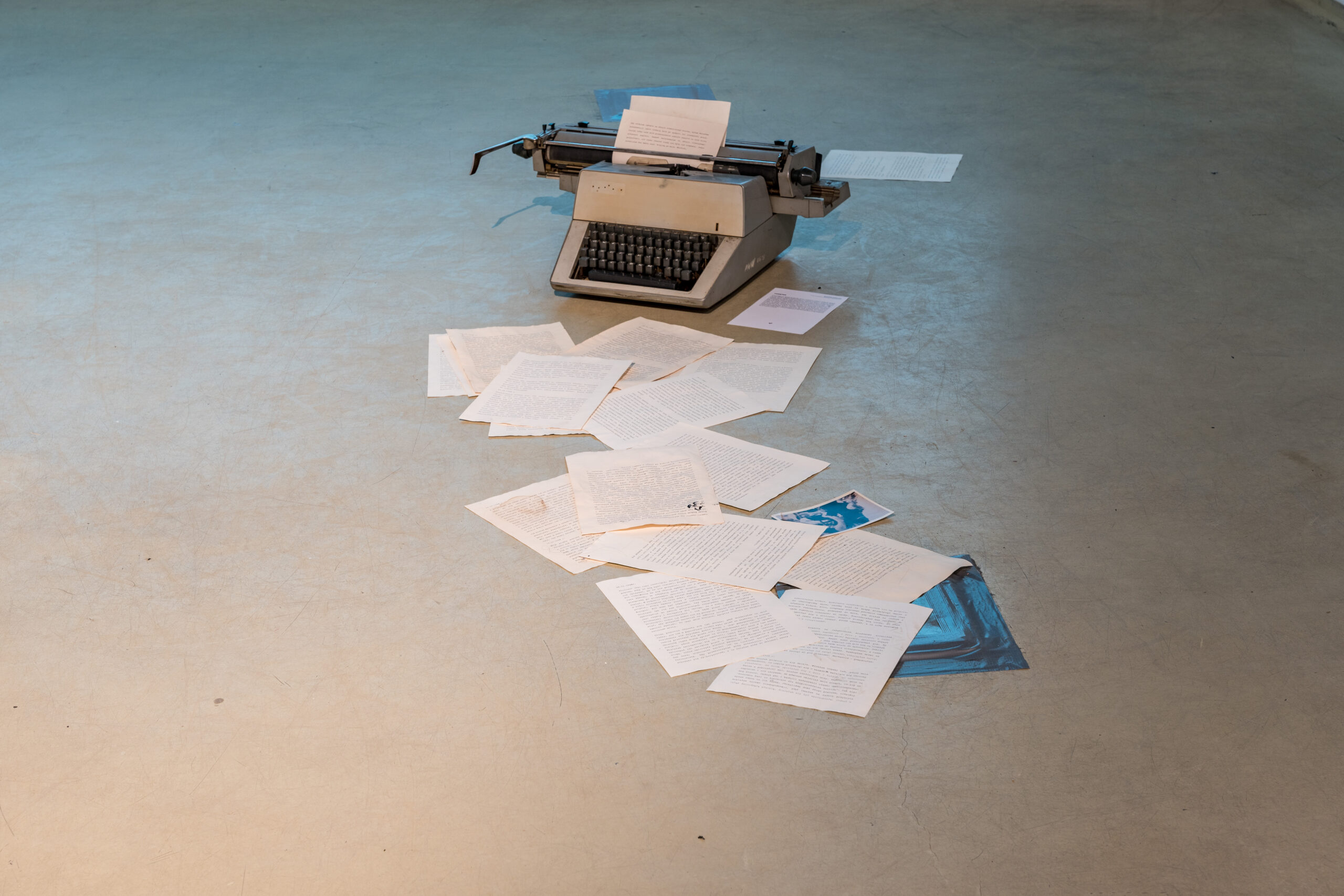 Na zdjęciu stara maszyna do pisania ustawiona na gładkiej posadzce, porozrzucane kartki papieru, stary maszynopis. 