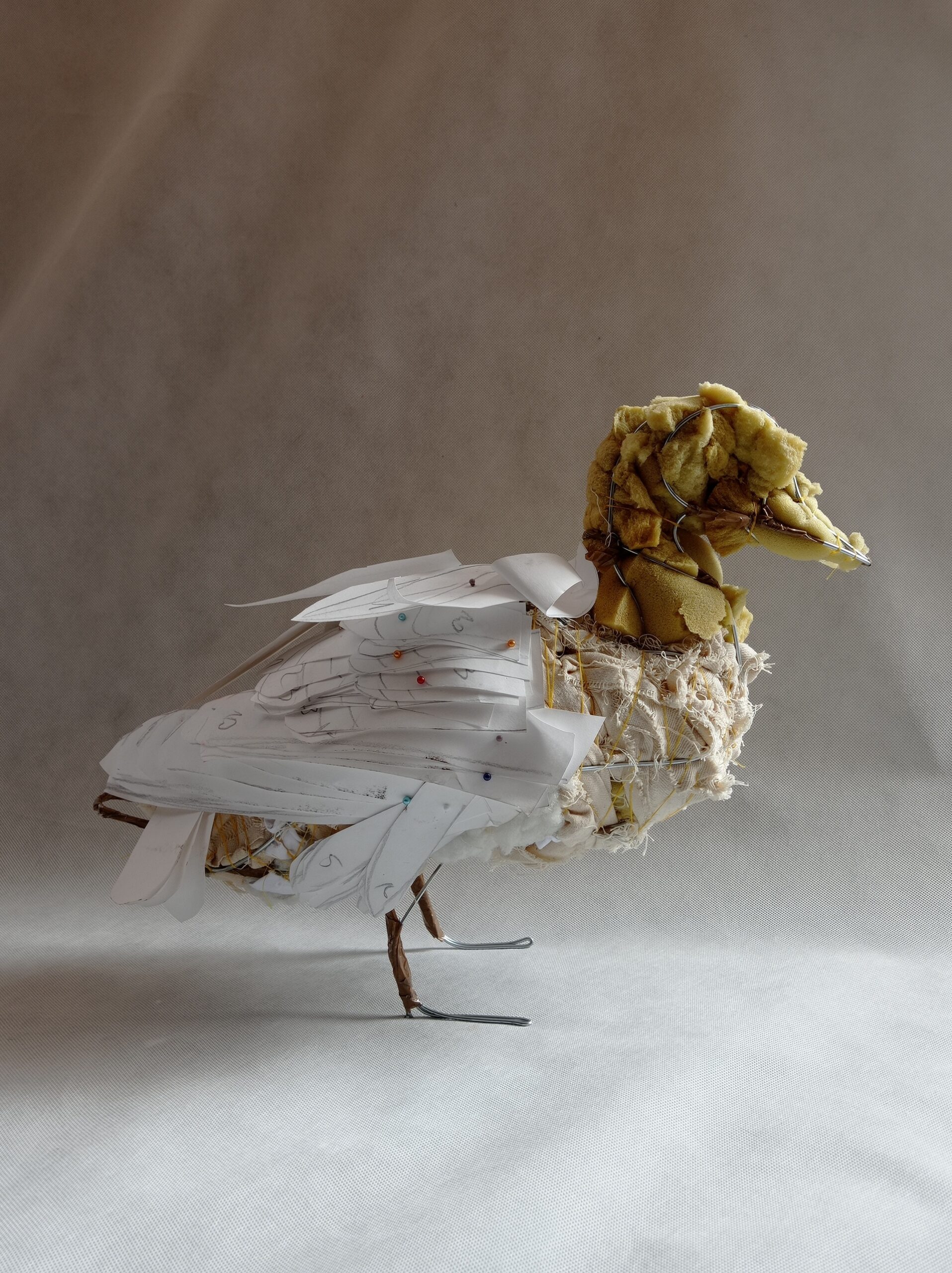 zdjęcie przedstawia rzeźbę kaczki, wykonaną z drutu, gąbki, materiału i papieru(elementy skrzydeł). Kaczka stoi na szarym tle, widok z boku