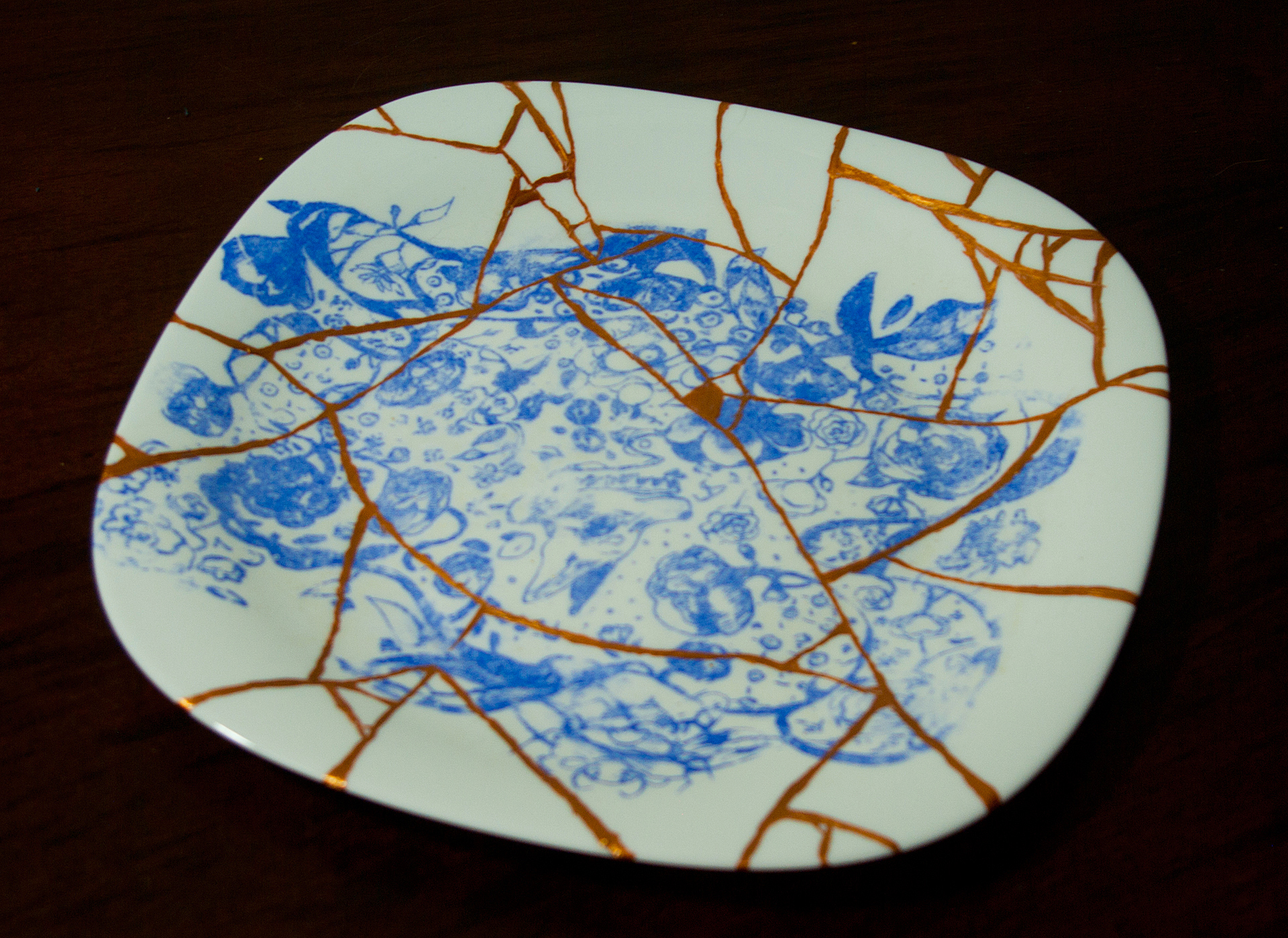Praca przedstawia trzy talerze, jeden z nich jest w całości, drugi rozbity, a trzeci sklejony. Na każdym z nich znajduje się nadruk w postaci niebieskiego, linearnego rysunku.