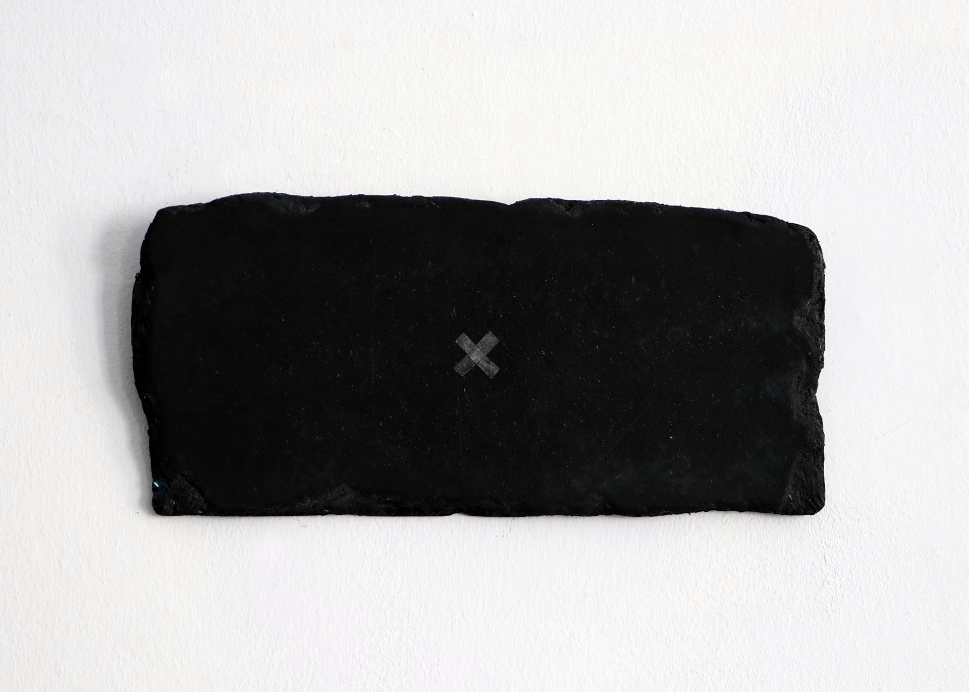 Zdjęcie przedstawia ciemną, drewnianą, prostokątną płytkę z naniesionym na nią niewielkim znakiem x, umieszczonym w jej środku