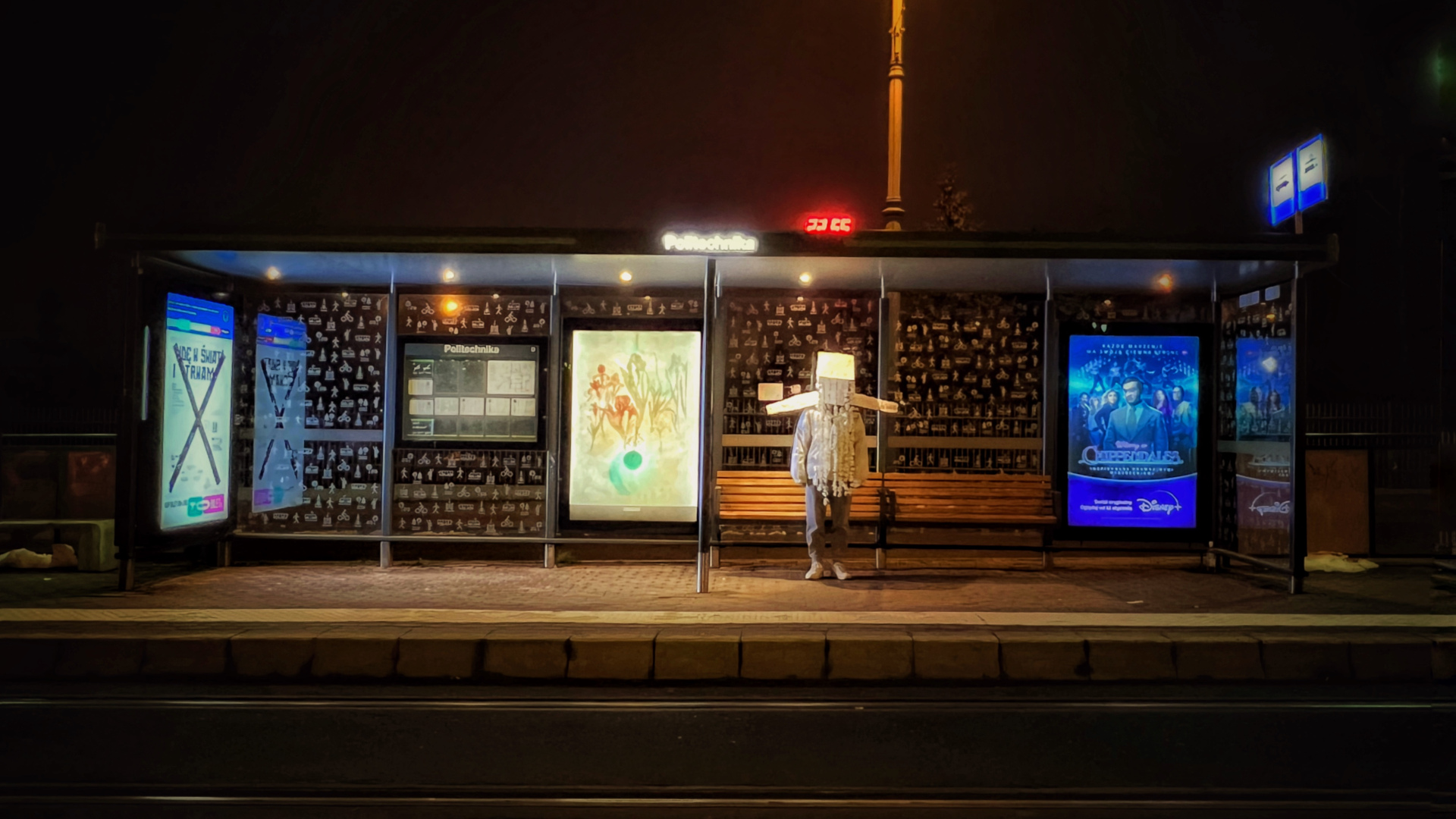 zdjęcie przedstawia postać ubraną w białą papierową koronę w formie hełmu. Postać stoi na przystanku autobusowym w nocy