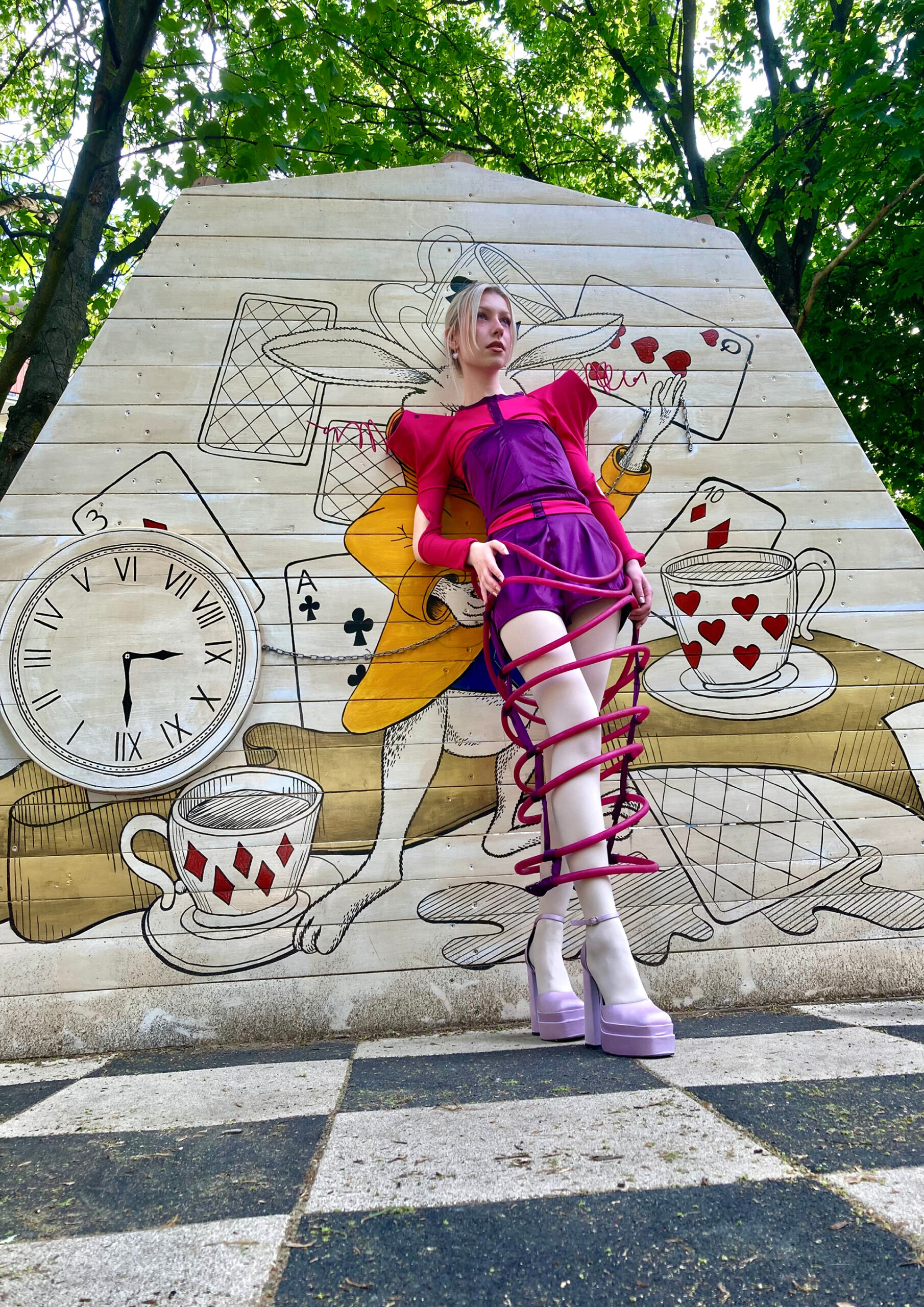 Kobieta w kolorowym, różowo-fioletowym kostiumie z obręczami wokół nóg i fiołkowych wysokich butach. Stoi na szachownicy, opiera się o ścianę z ilustracją przedstawiającą królika, zegar i karty, za ścianą widoczne są korony drzew.