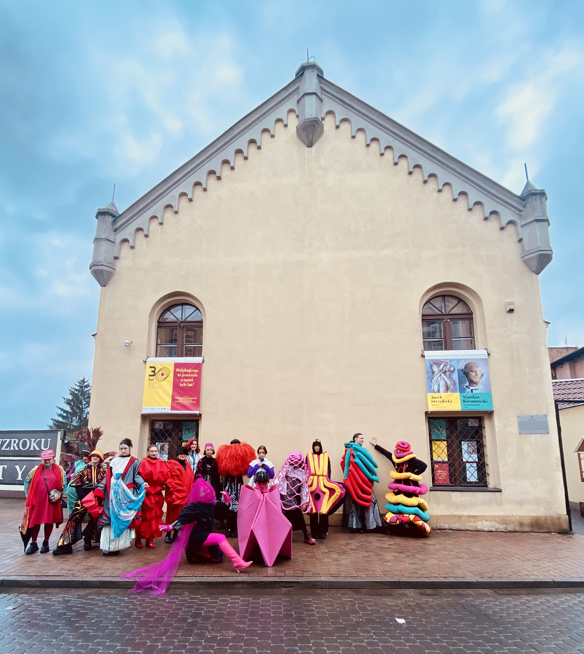Grupa osób performujących w wielokolorowych kostiumach na tle budynku Galerii Sztuki w Mosinie.