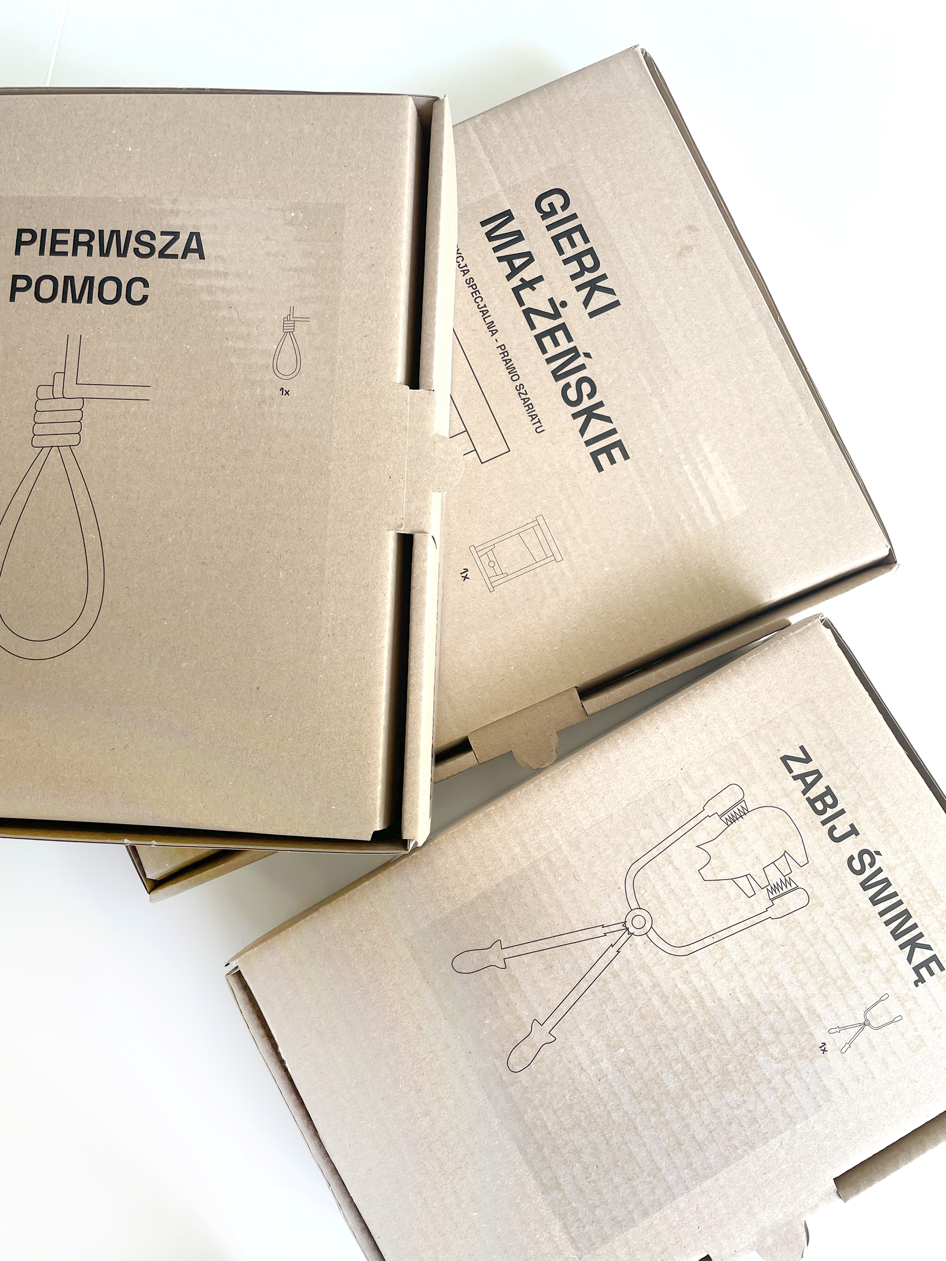 Zdjęcie trzech zamkniętych, leżących jeden na drugim kartonów z przezroczystymi grafikami w stylu firmy „Ikea”. Obrazki i napisy przedstawiają – od lewej: wisielczy sznur - „Pierwsza pomoc”, „Gierki małżeńskie” oraz narzędzie trzymające małą świnkę - „Zabij świnkę”.