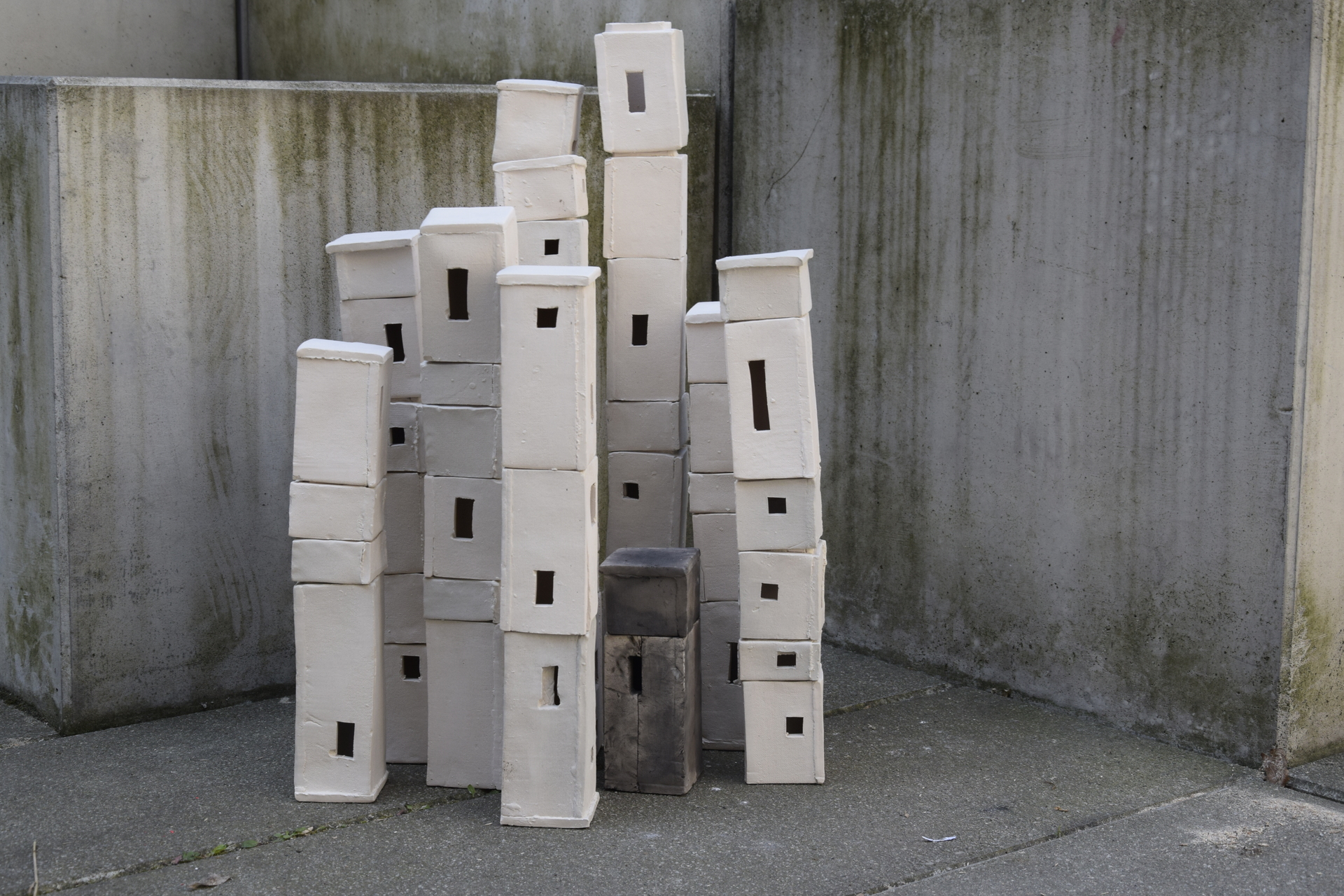 Grupa obiektów ceramicznych, wykonanych z jasnej, beżowej gliny. Prostopadłościenne elementy, ustawione na sobie, układają się w kształt wież. Część ma wycięte prostokątne otwory. Na betonowym tle